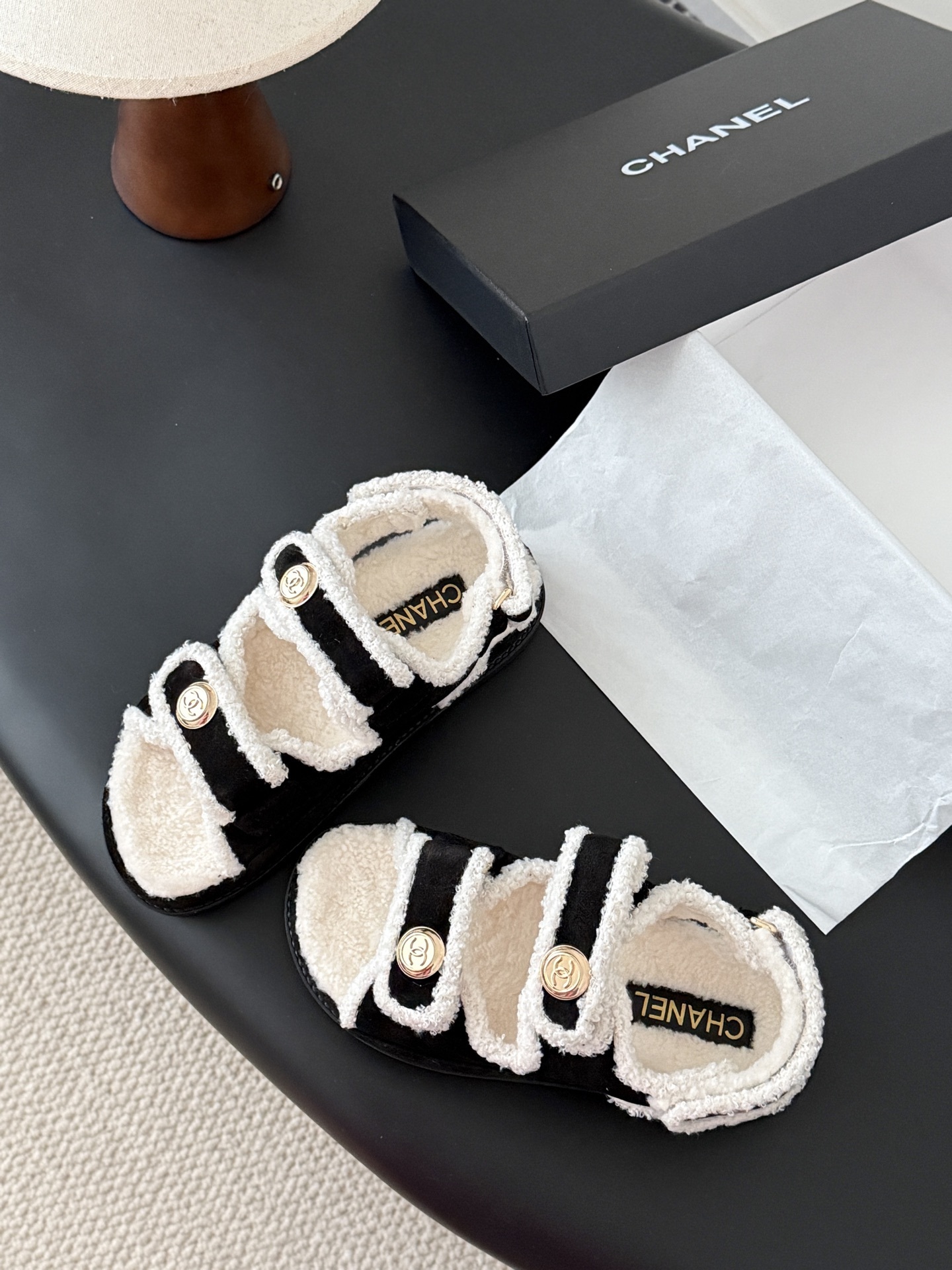 Chanel black and white shearling dad sandals with gold CC logo buttons on a black table next to a Chanel box - Hình 6