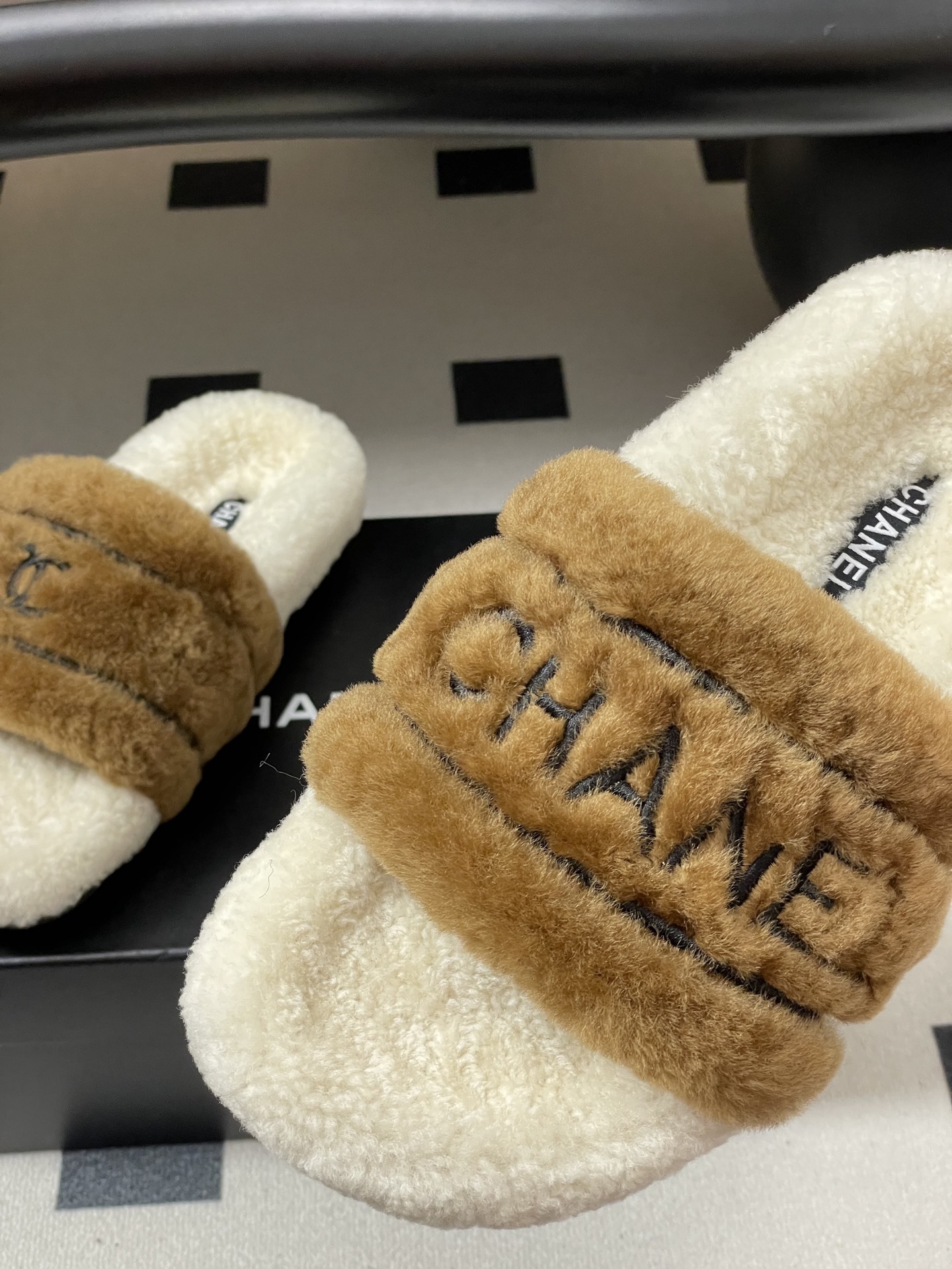 A pair of Chanel branded faux fur slides in brown and white shearling on a black surface. - Hình 9