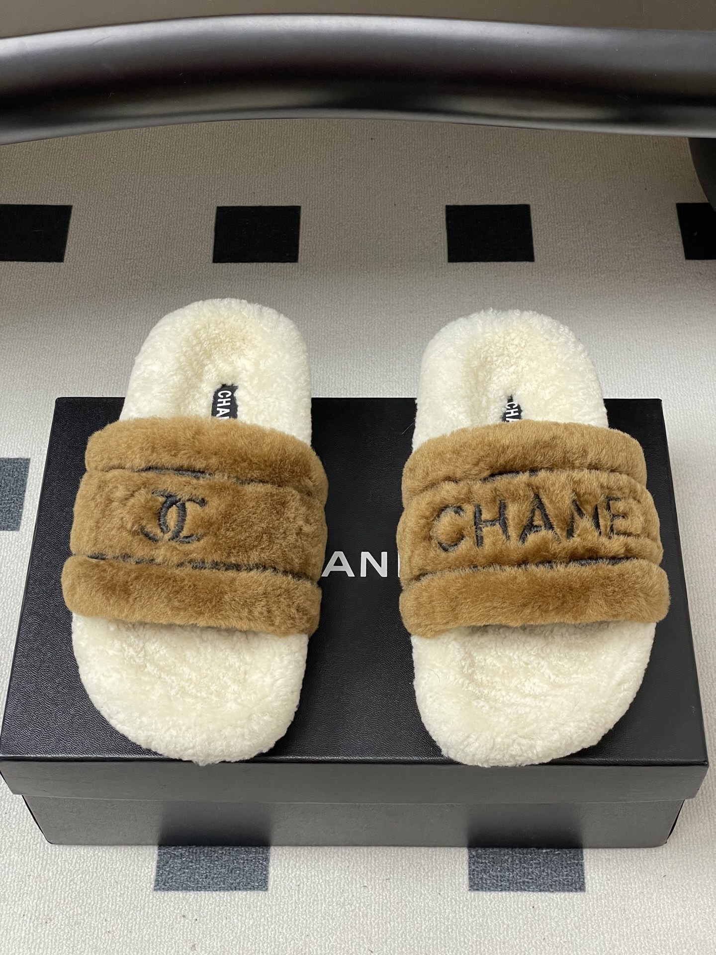 A pair of Chanel branded faux fur slides in brown and white shearling on a black surface. - Hình 5