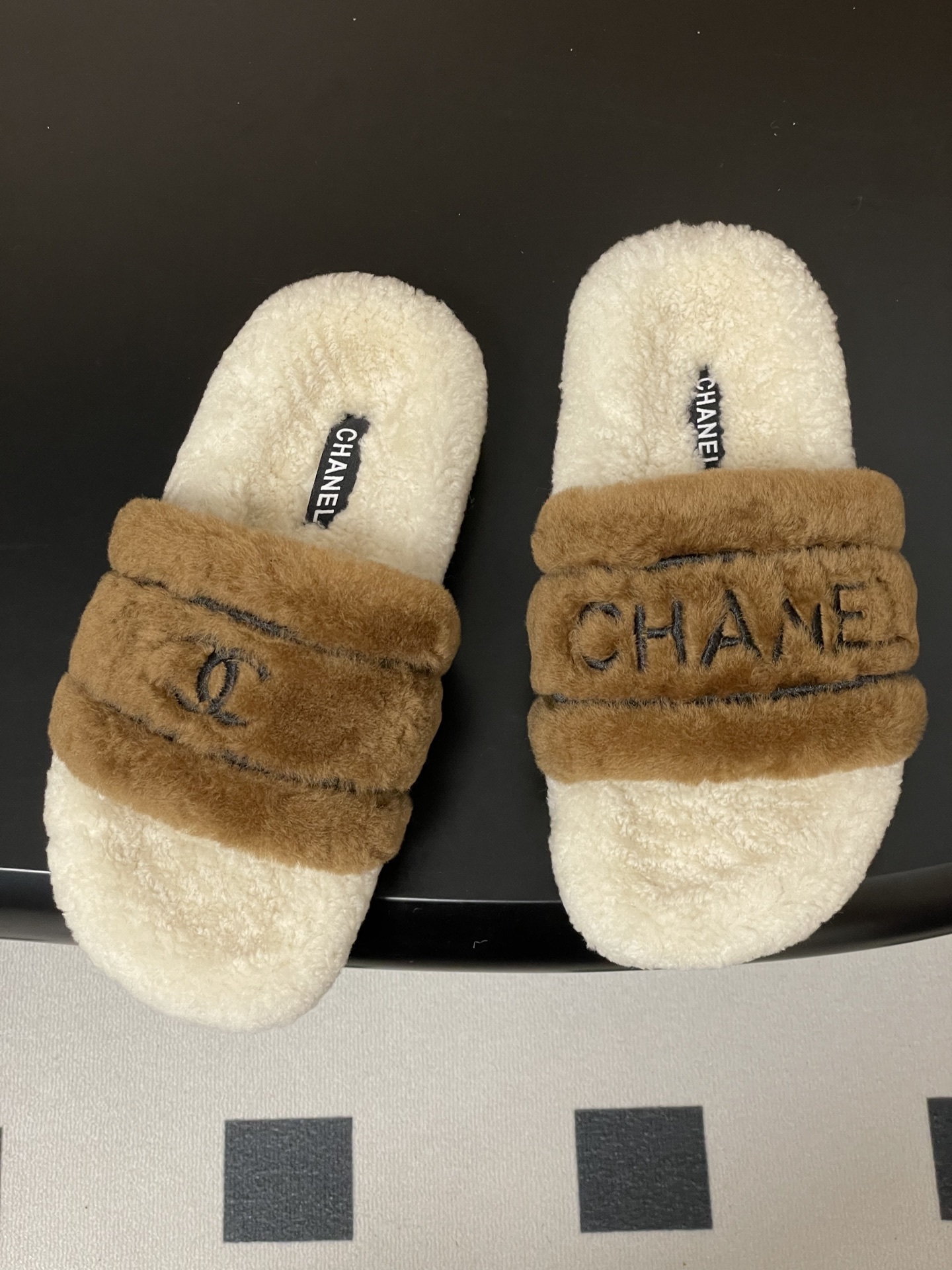 A pair of Chanel branded faux fur slides in brown and white shearling on a black surface. - Hình 2
