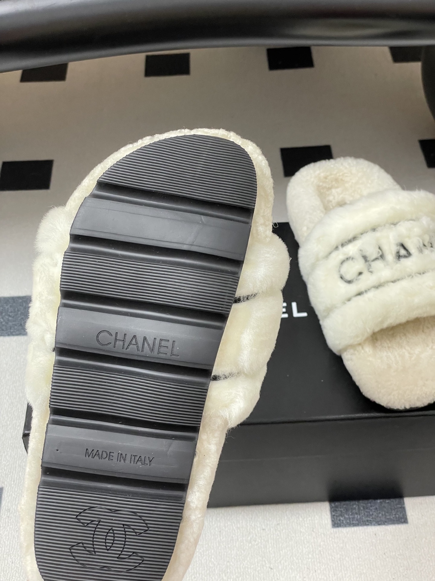 Pair of luxury white Chanel fuzzy shearling slides with black logo embroidery displayed on a black table. - Hình 9