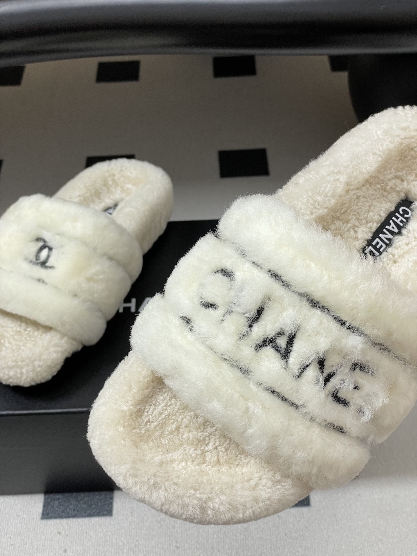 Pair of luxury white Chanel fuzzy shearling slides with black logo embroidery displayed on a black table. - Hình 8