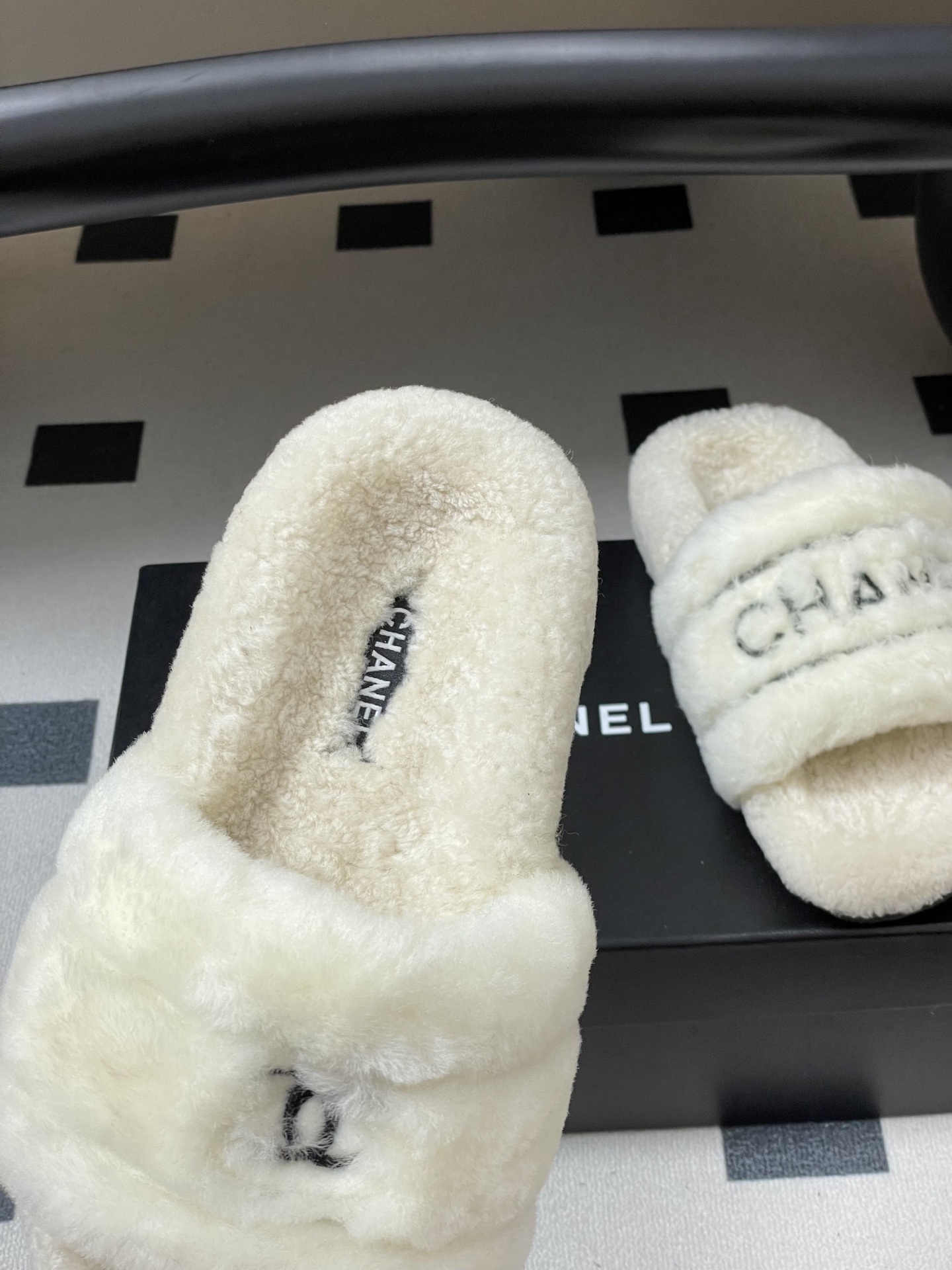 Pair of luxury white Chanel fuzzy shearling slides with black logo embroidery displayed on a black table. - Hình 7