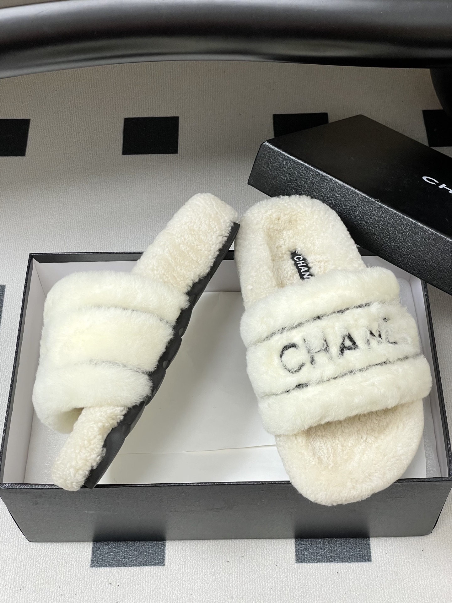 Pair of luxury white Chanel fuzzy shearling slides with black logo embroidery displayed on a black table. - Hình 6