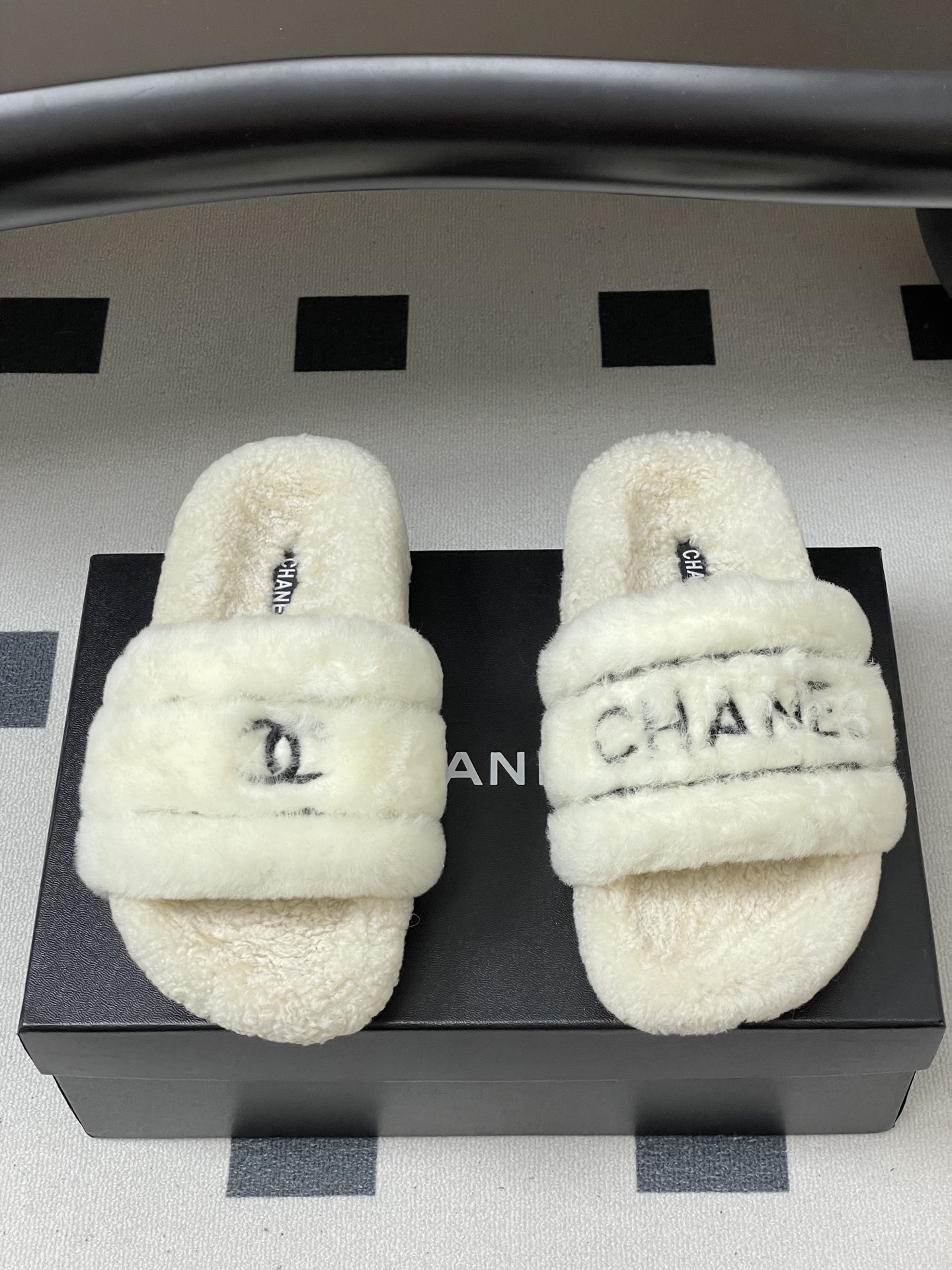 Pair of luxury white Chanel fuzzy shearling slides with black logo embroidery displayed on a black table. - Hình 5