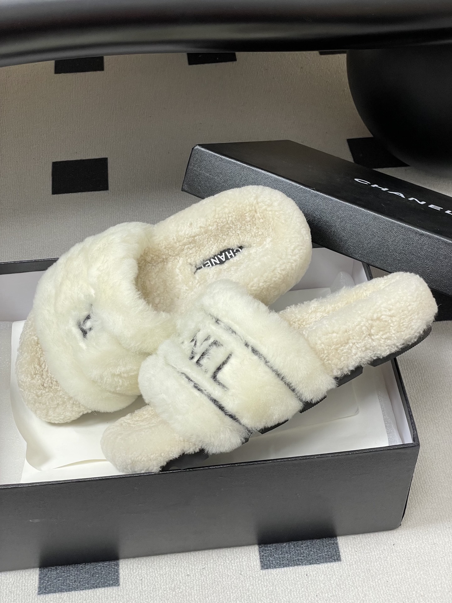Pair of luxury white Chanel fuzzy shearling slides with black logo embroidery displayed on a black table. - Hình 4