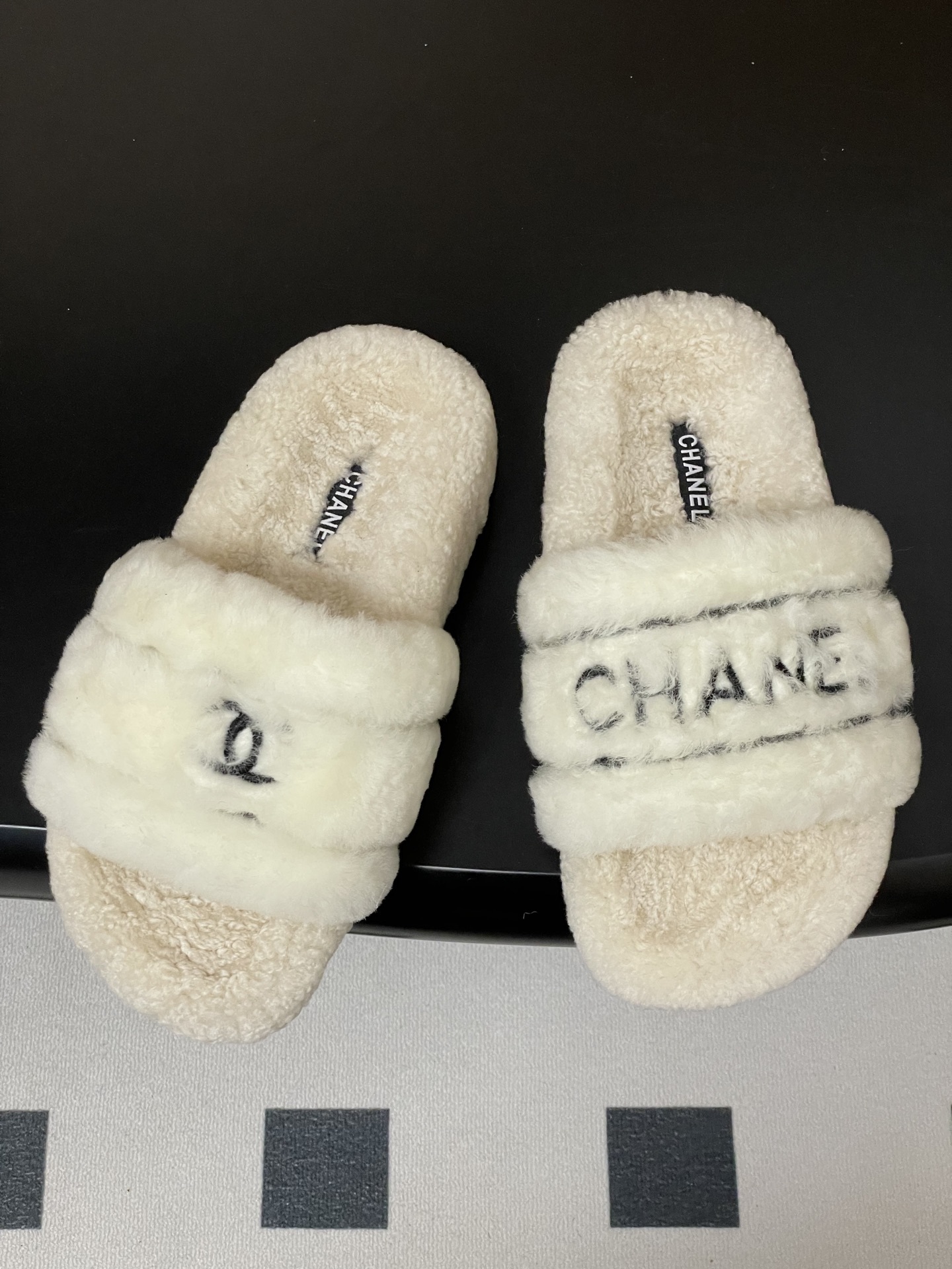 Pair of luxury white Chanel fuzzy shearling slides with black logo embroidery displayed on a black table. - Hình 2