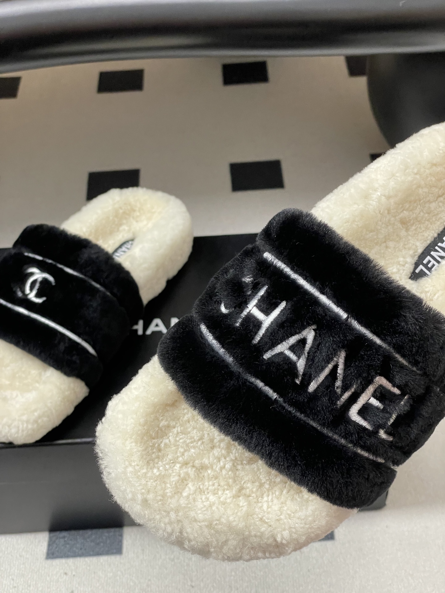 A pair of black and white Chanel shearling slides with silver logo embroidery on a black surface - Hình 9