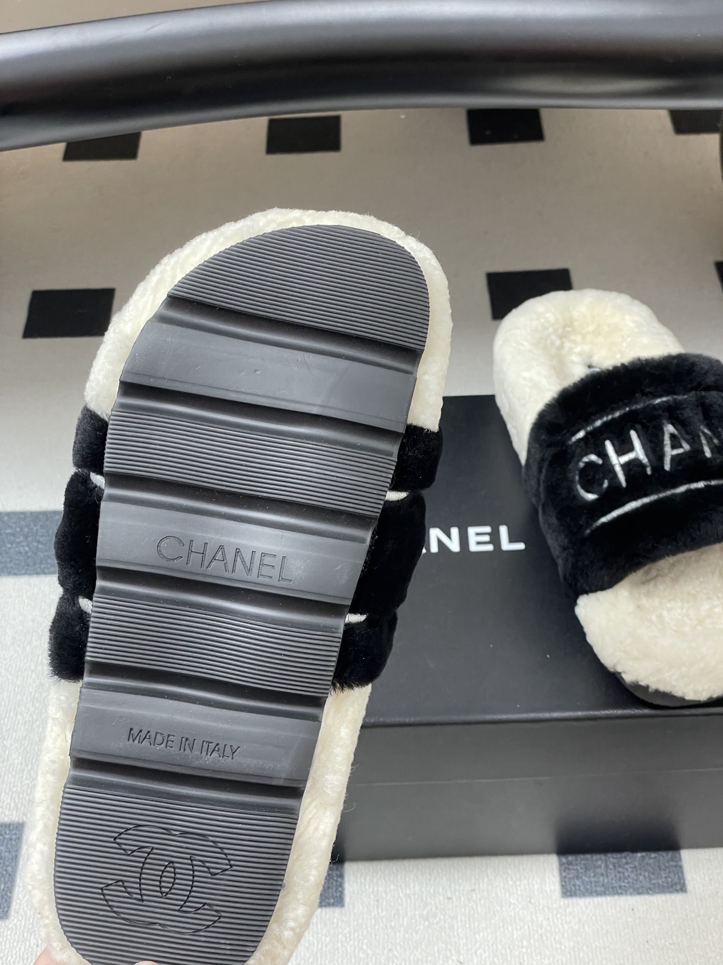 A pair of black and white Chanel shearling slides with silver logo embroidery on a black surface - Hình 8