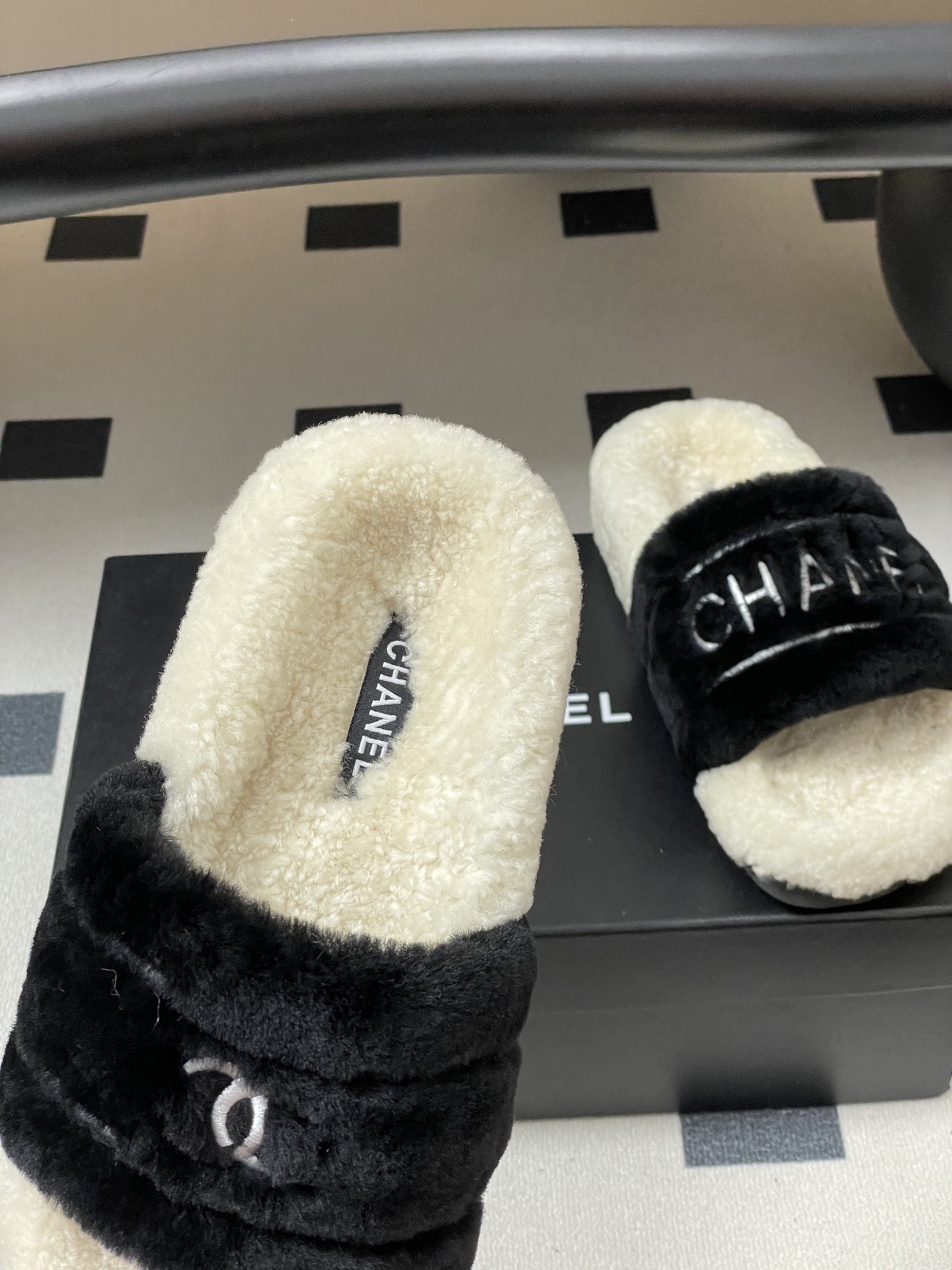 A pair of black and white Chanel shearling slides with silver logo embroidery on a black surface - Hình 7