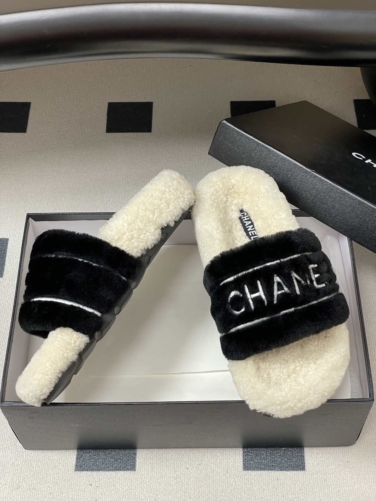 A pair of black and white Chanel shearling slides with silver logo embroidery on a black surface - Hình 6
