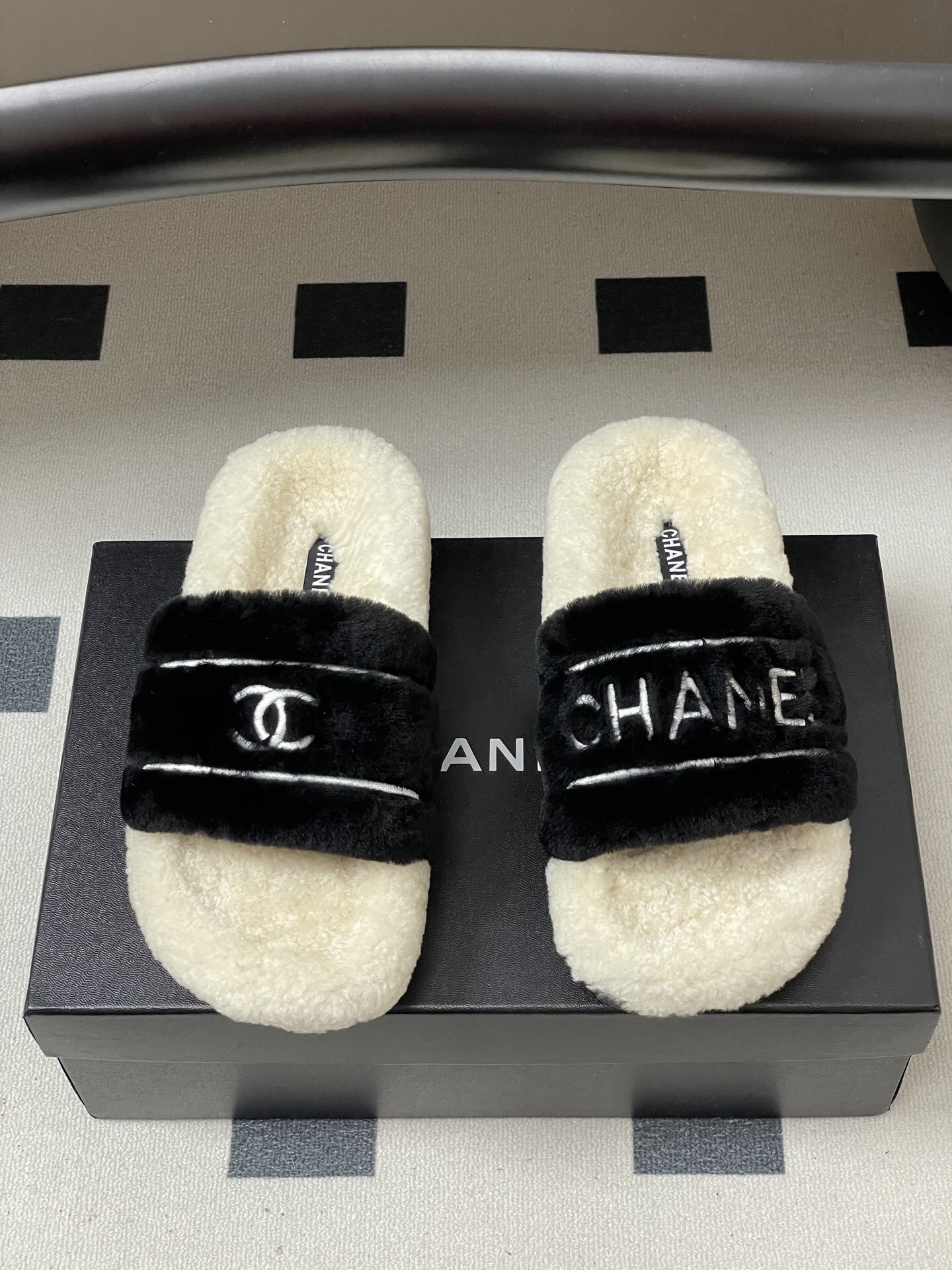A pair of black and white Chanel shearling slides with silver logo embroidery on a black surface - Hình 5