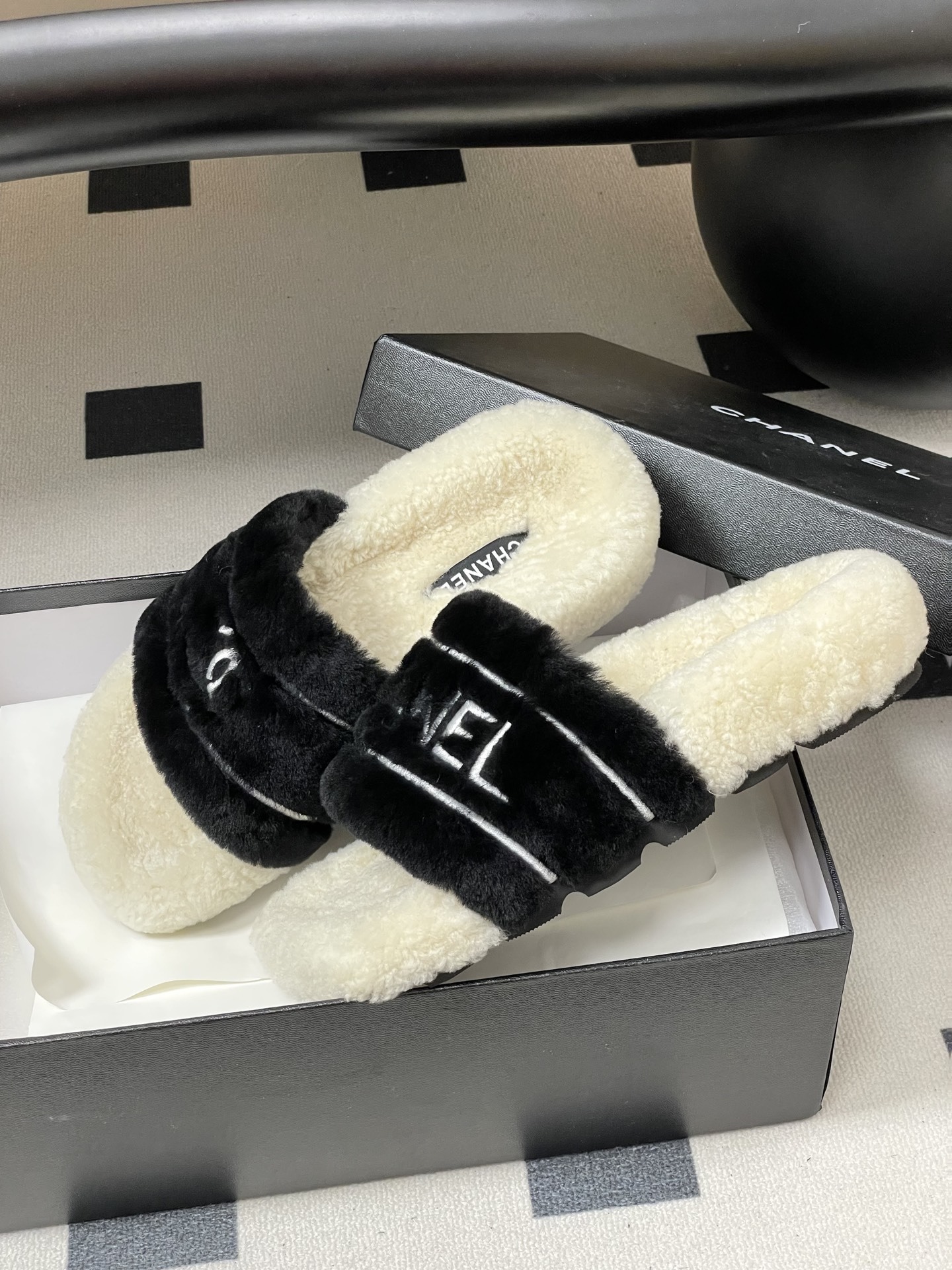 A pair of black and white Chanel shearling slides with silver logo embroidery on a black surface - Hình 4