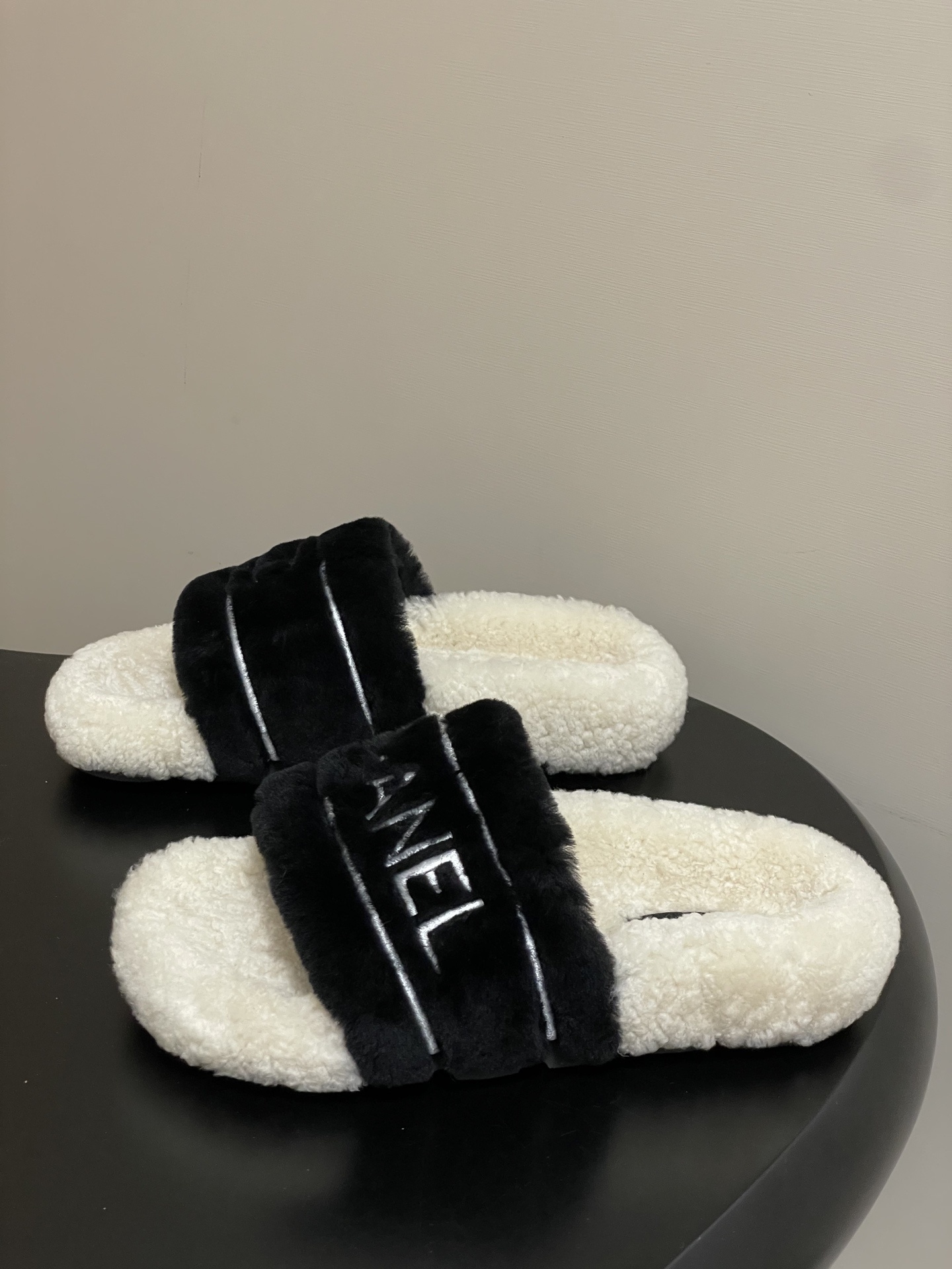 A pair of black and white Chanel shearling slides with silver logo embroidery on a black surface - Hình 3
