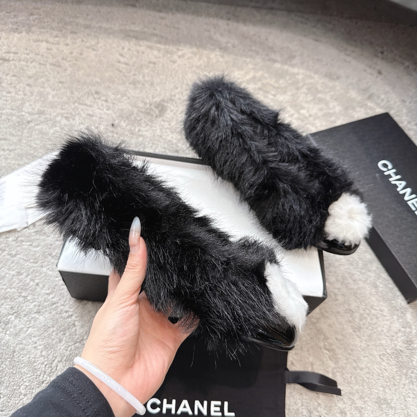 Top view of black Chanel fur loafers with white pom-poms on a grey carpet - Hình 3
