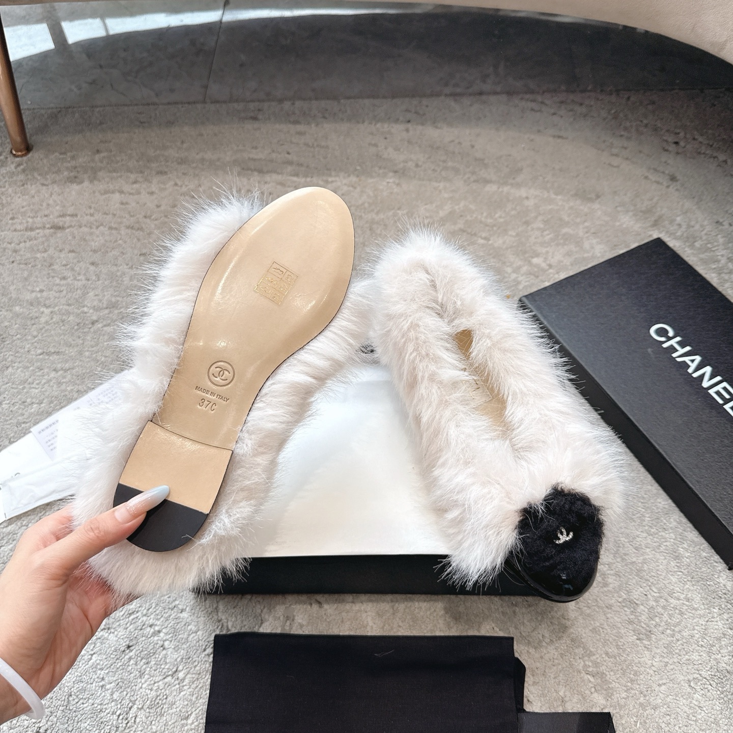 A pair of luxury white faux fur ballerina flats with black toe caps and silver CC logos, presented in a designer box. - Hình 9