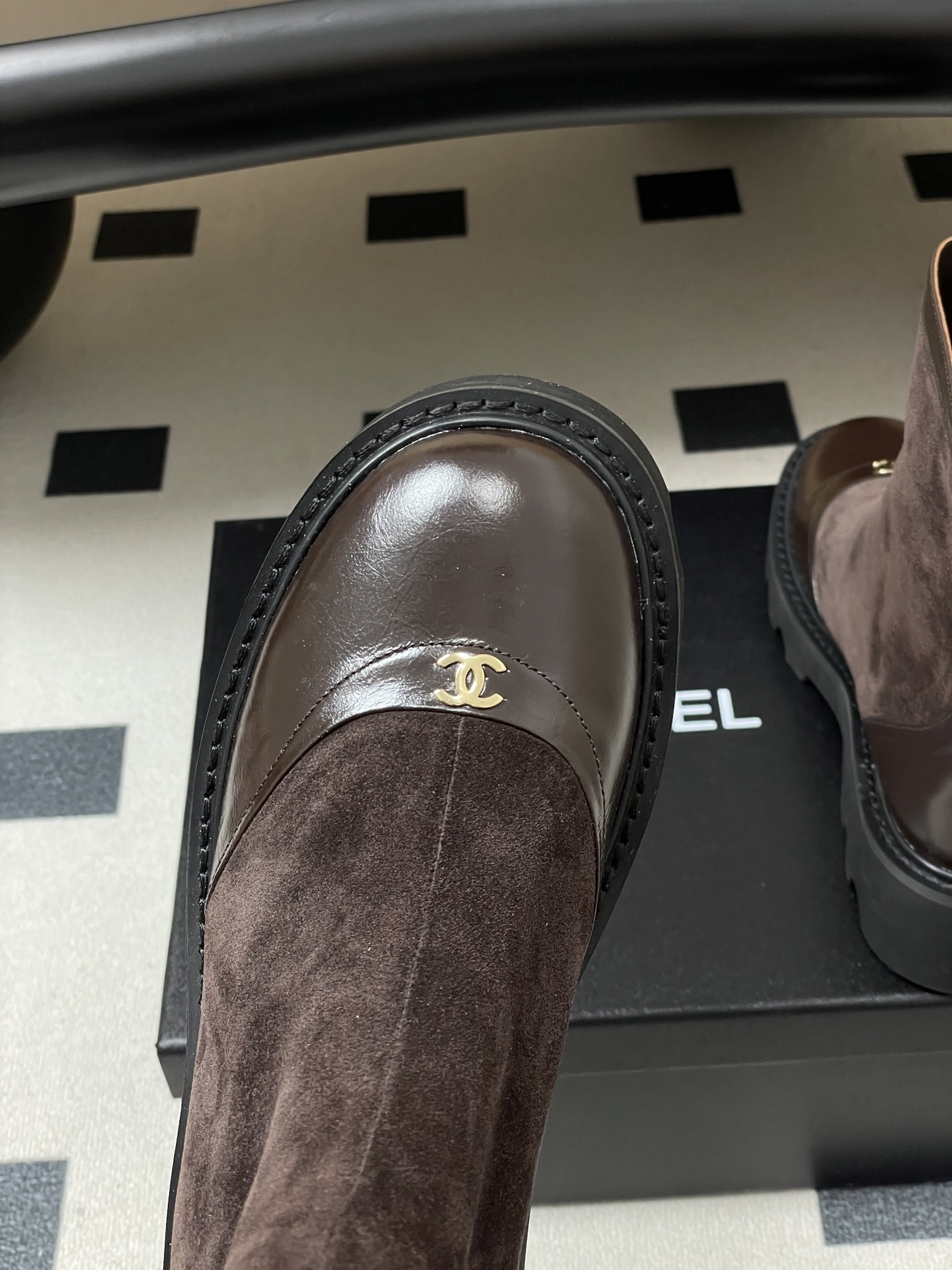 A pair of Chanel dark brown suede and leather ankle boots with gold CC logos and thick black lug soles on a black table. - Hình 7