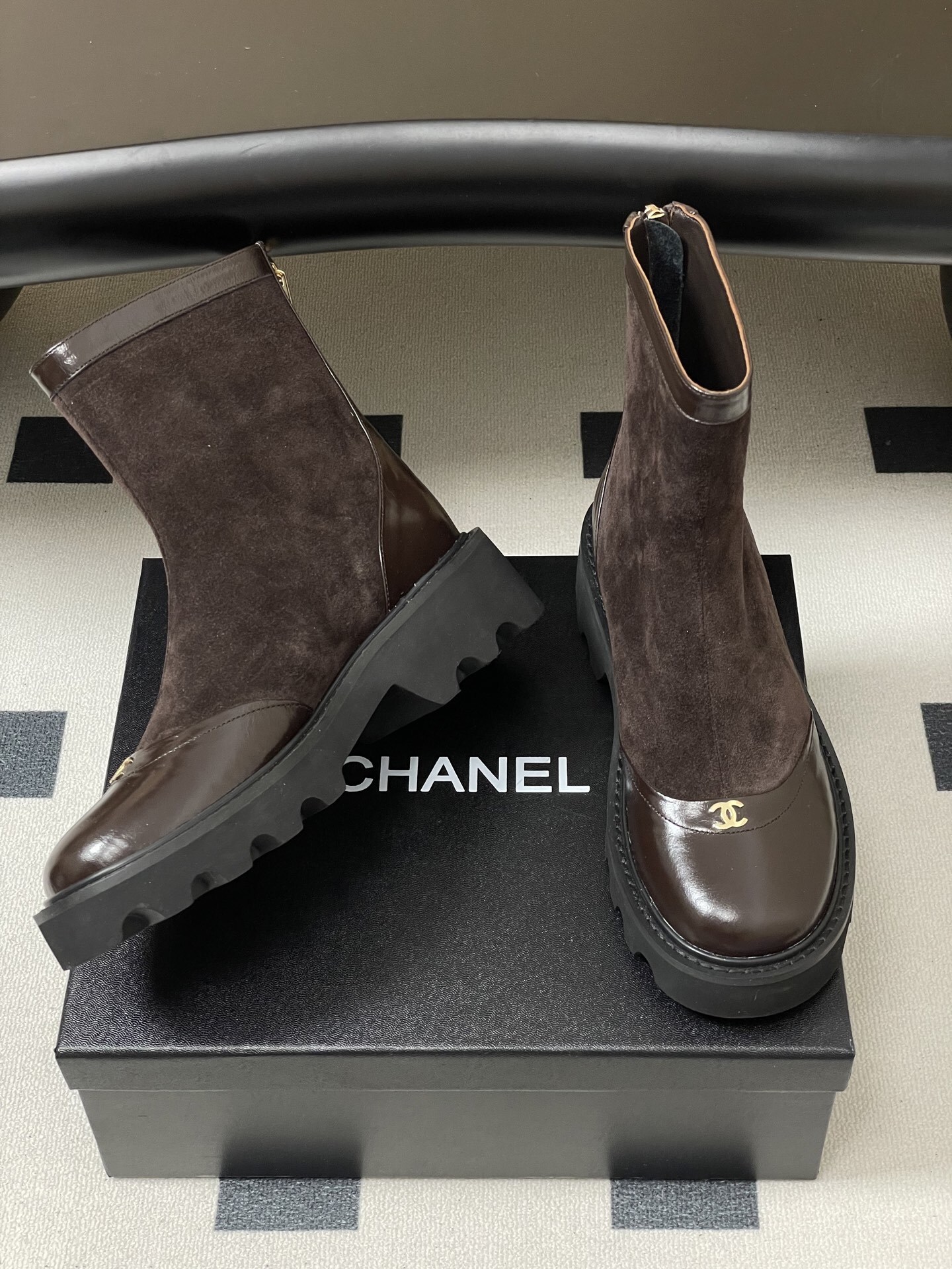 A pair of Chanel dark brown suede and leather ankle boots with gold CC logos and thick black lug soles on a black table. - Hình 5