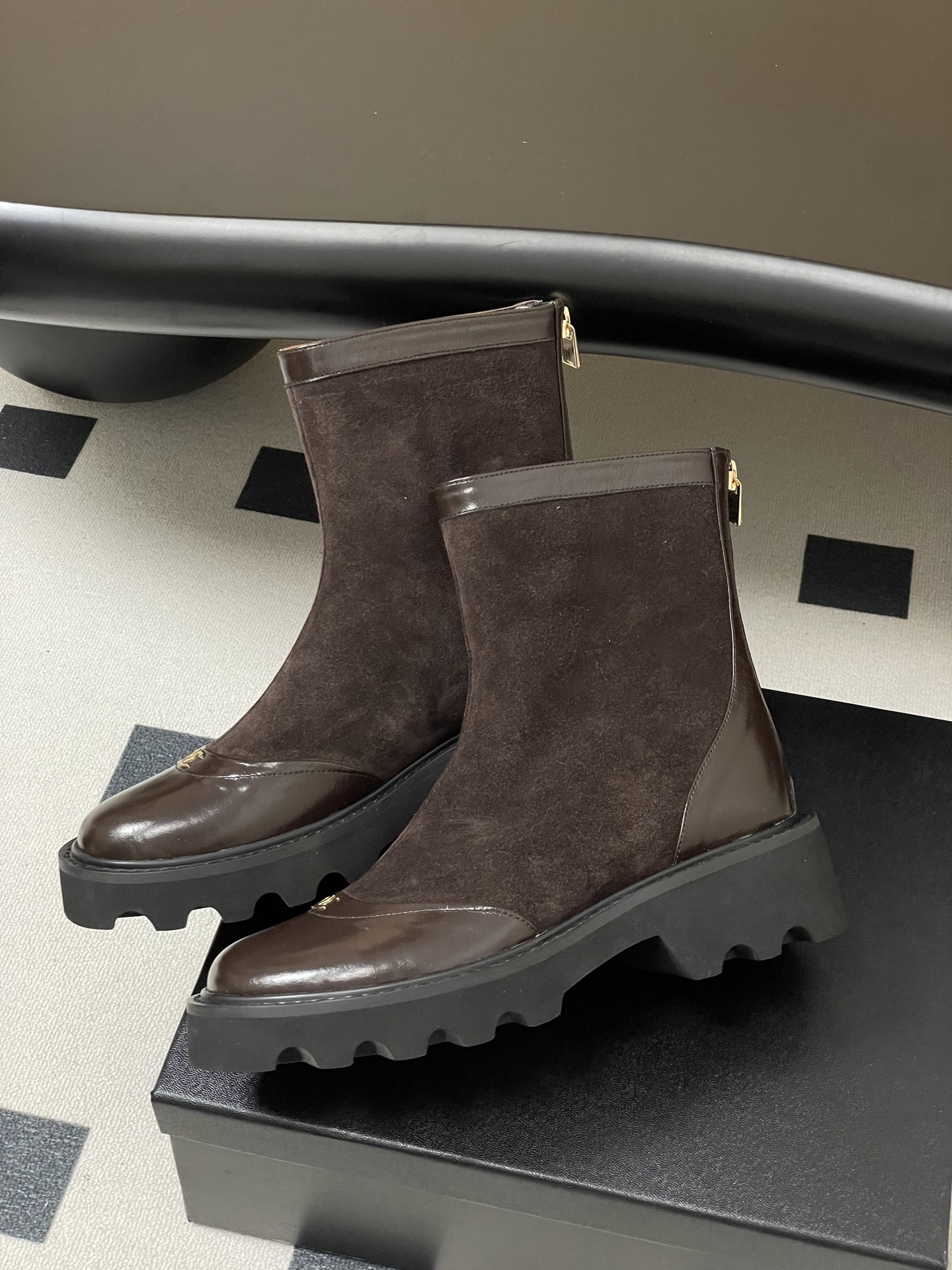 A pair of Chanel dark brown suede and leather ankle boots with gold CC logos and thick black lug soles on a black table. - Hình 4