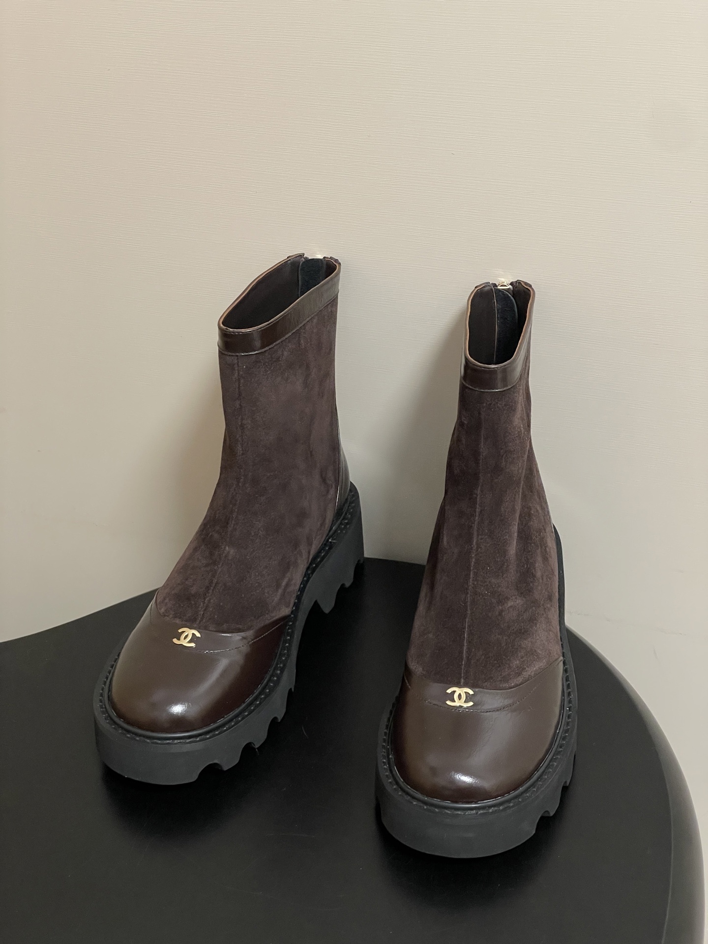 A pair of Chanel dark brown suede and leather ankle boots with gold CC logos and thick black lug soles on a black table. - Hình 2