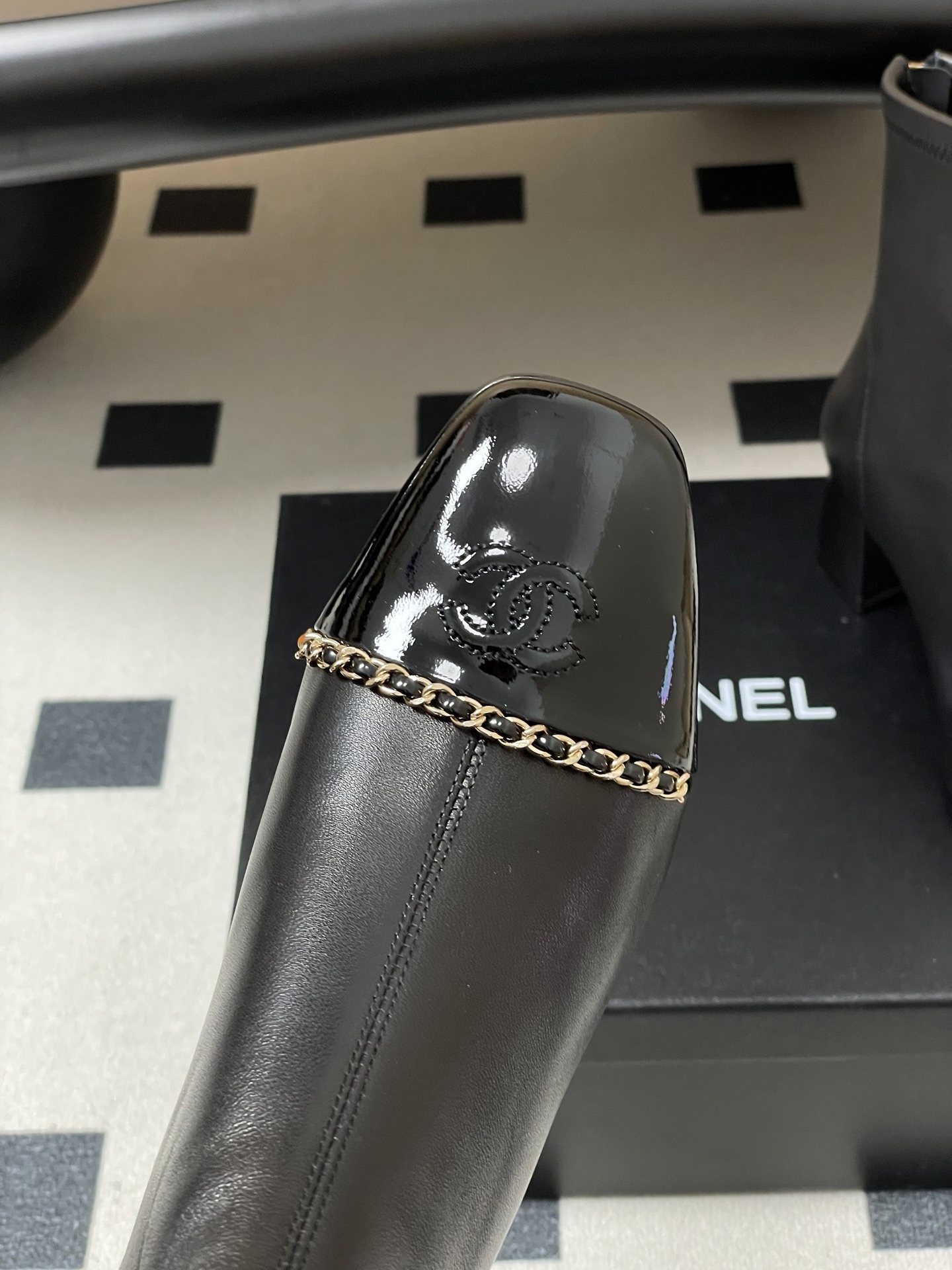 Elegant black leather ankle boots with gold chain detail and patent leather square toe cap on a black table. - Hình 5