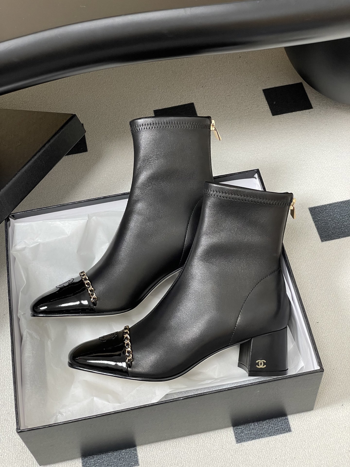 Elegant black leather ankle boots with gold chain detail and patent leather square toe cap on a black table. - Hình 4