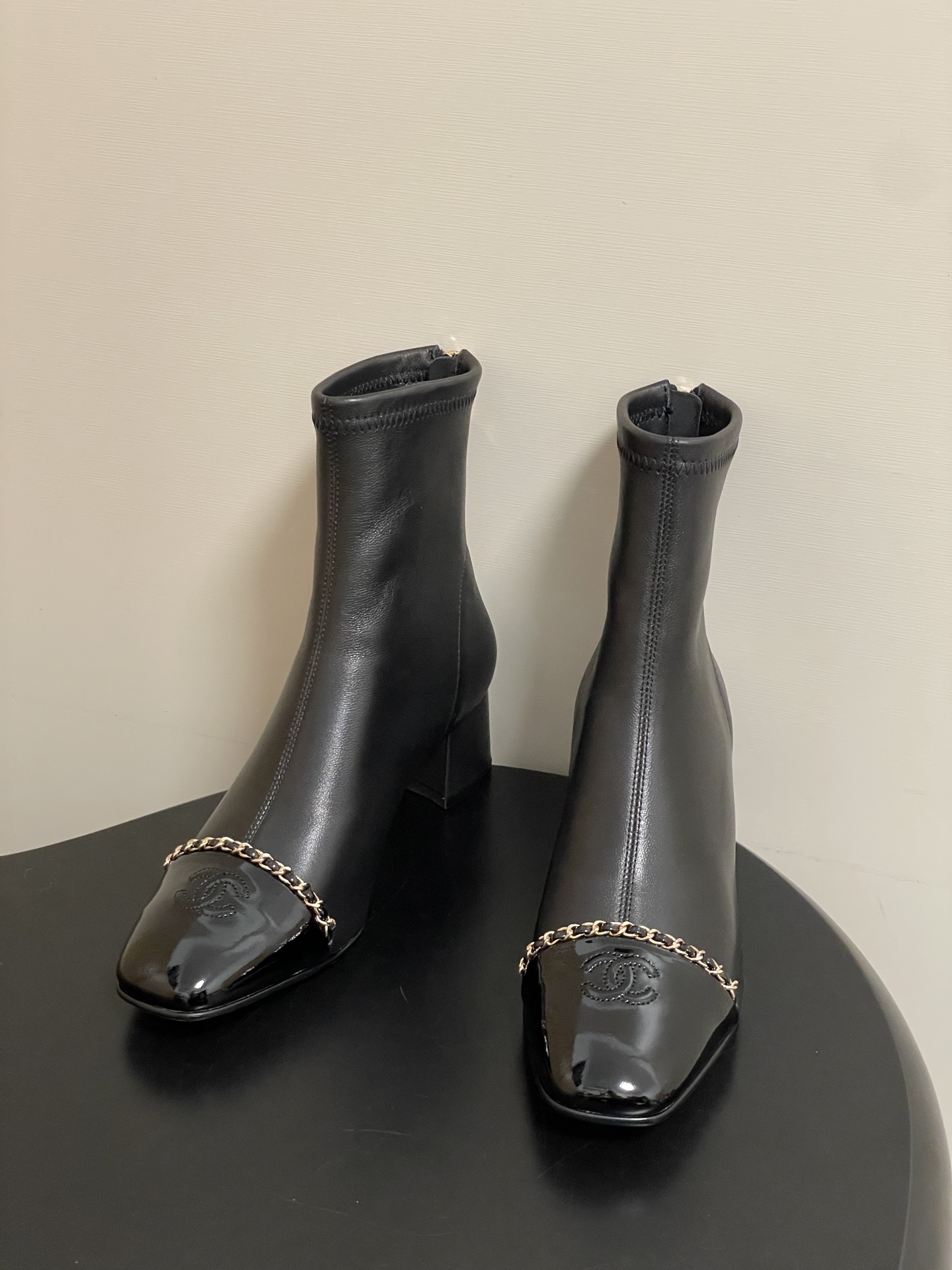 Elegant black leather ankle boots with gold chain detail and patent leather square toe cap on a black table. - Hình 2