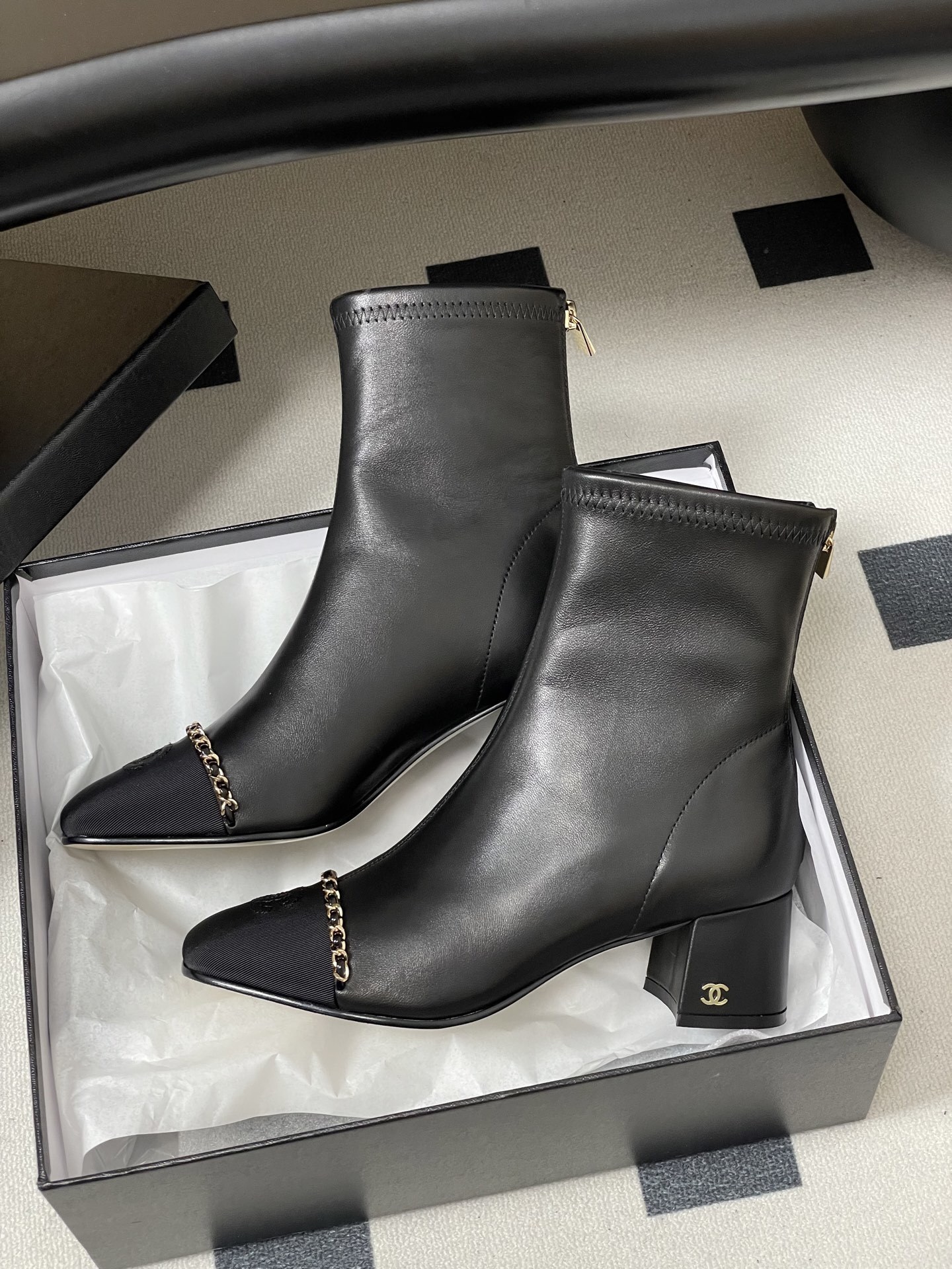 Pair of Chanel black leather ankle boots with gold chain detail and block heels on a black table - Hình 5