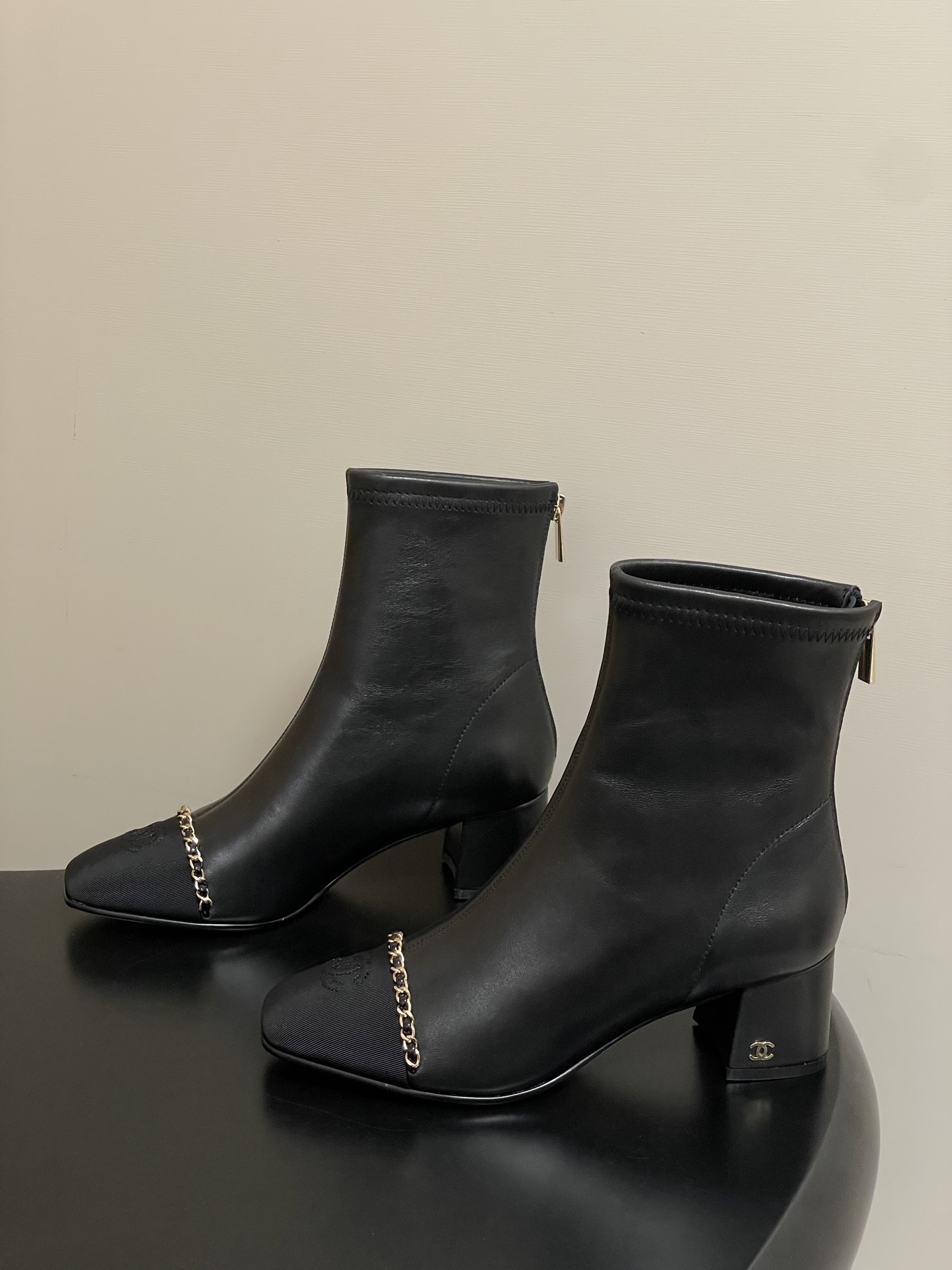 Pair of Chanel black leather ankle boots with gold chain detail and block heels on a black table - Hình 4