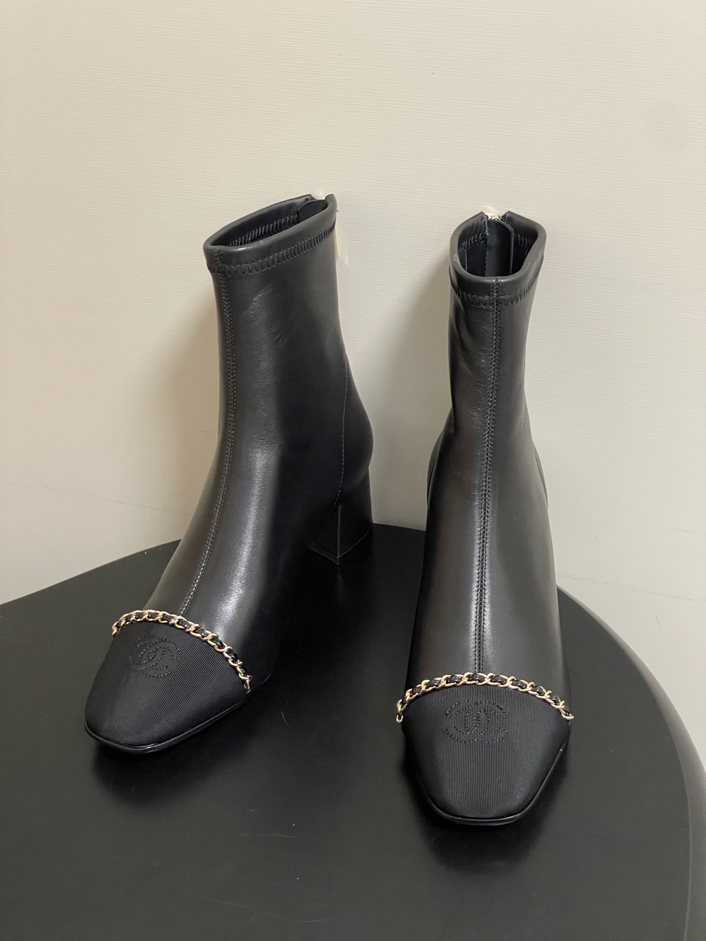 Pair of Chanel black leather ankle boots with gold chain detail and block heels on a black table - Hình 3