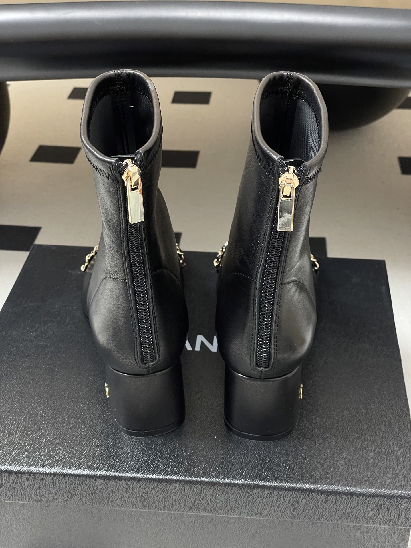 Pair of Chanel black leather ankle boots with gold chain detail and block heels on a black table - Hình 2