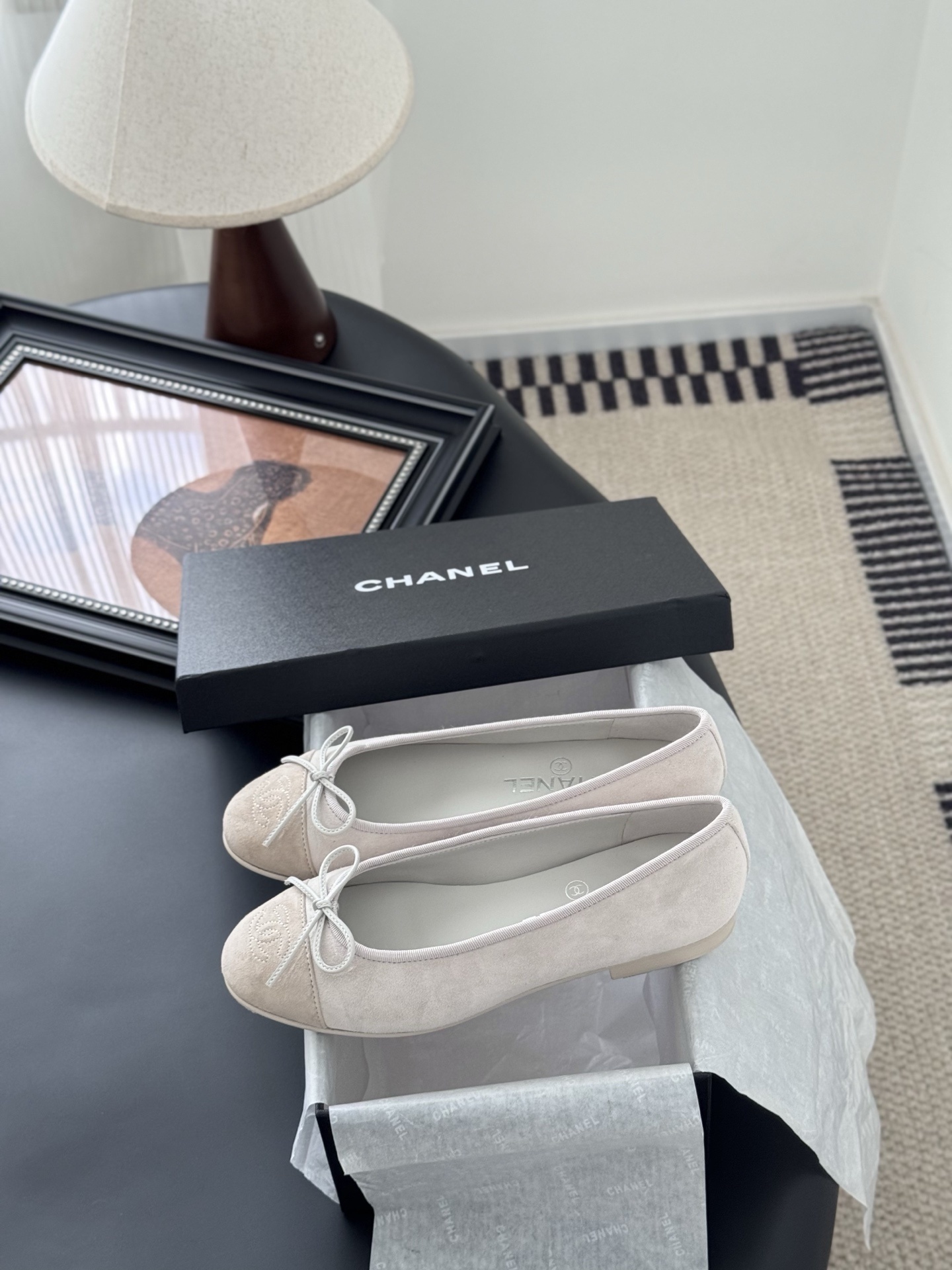 Pair of Chanel beige suede ballet flats with CC logo on the toe cap and white bow detail, placed next to a black Chanel box. - Hình 9