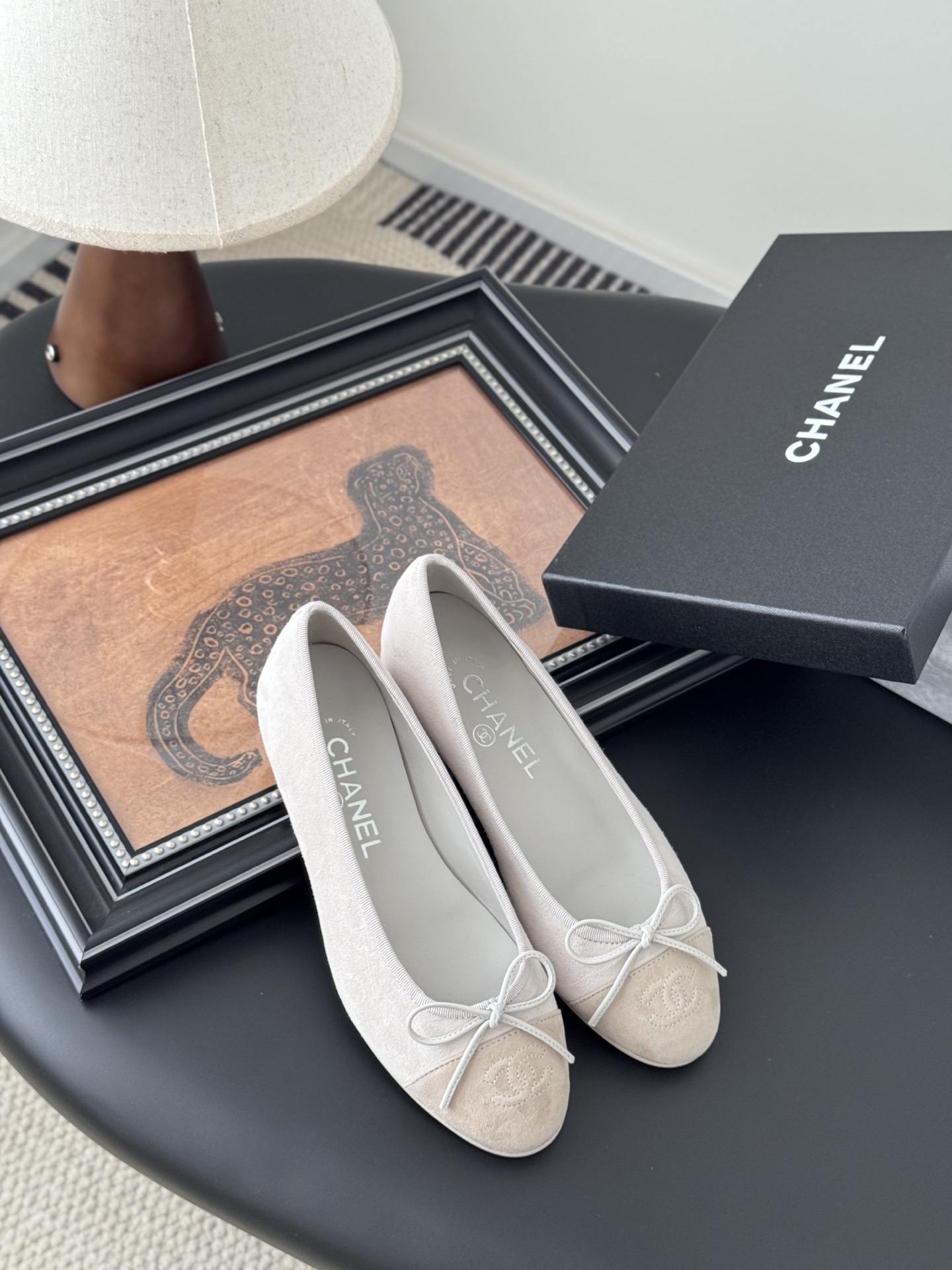 Pair of Chanel beige suede ballet flats with CC logo on the toe cap and white bow detail, placed next to a black Chanel box. - Hình 7