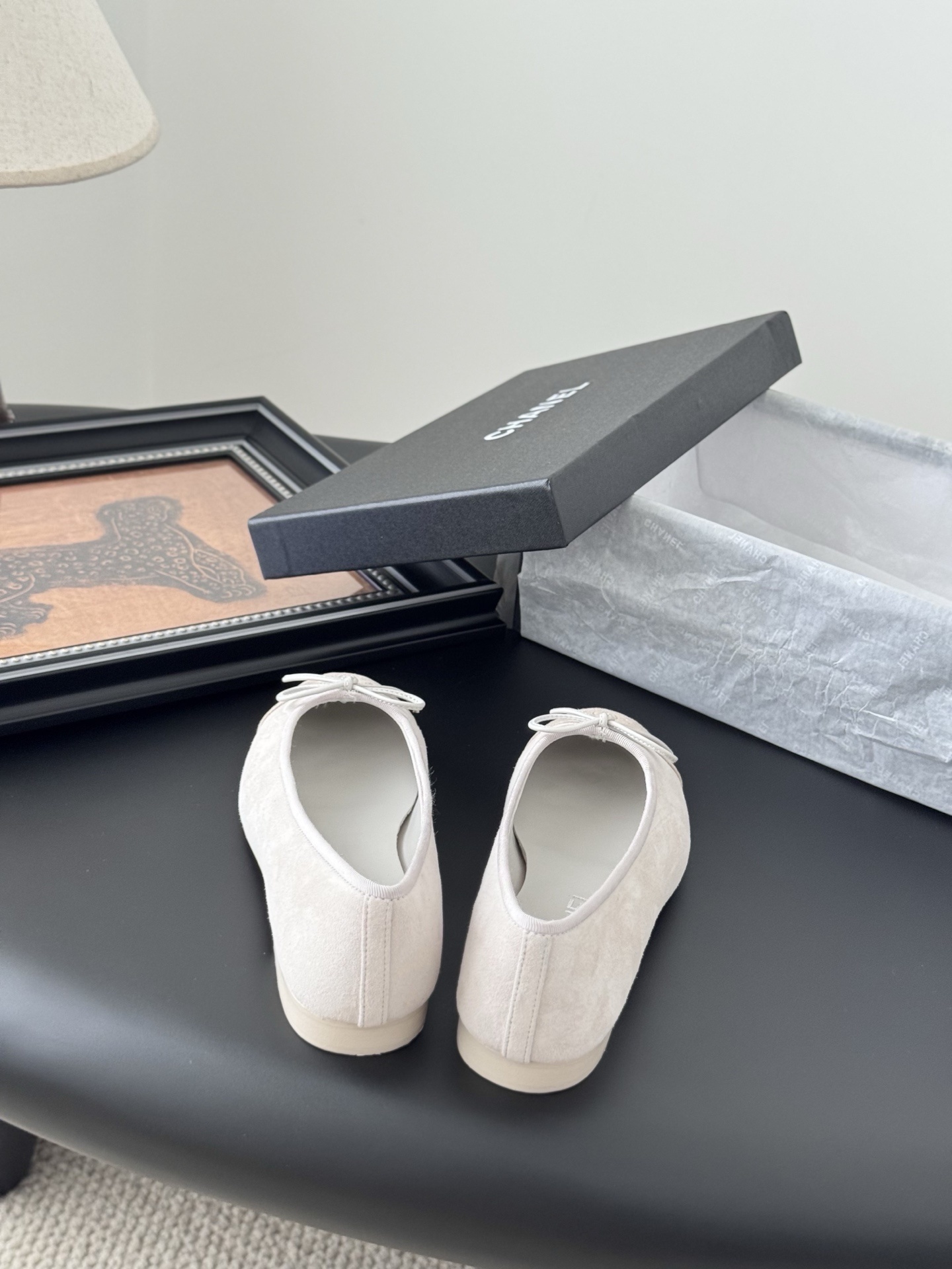 Pair of Chanel beige suede ballet flats with CC logo on the toe cap and white bow detail, placed next to a black Chanel box. - Hình 6