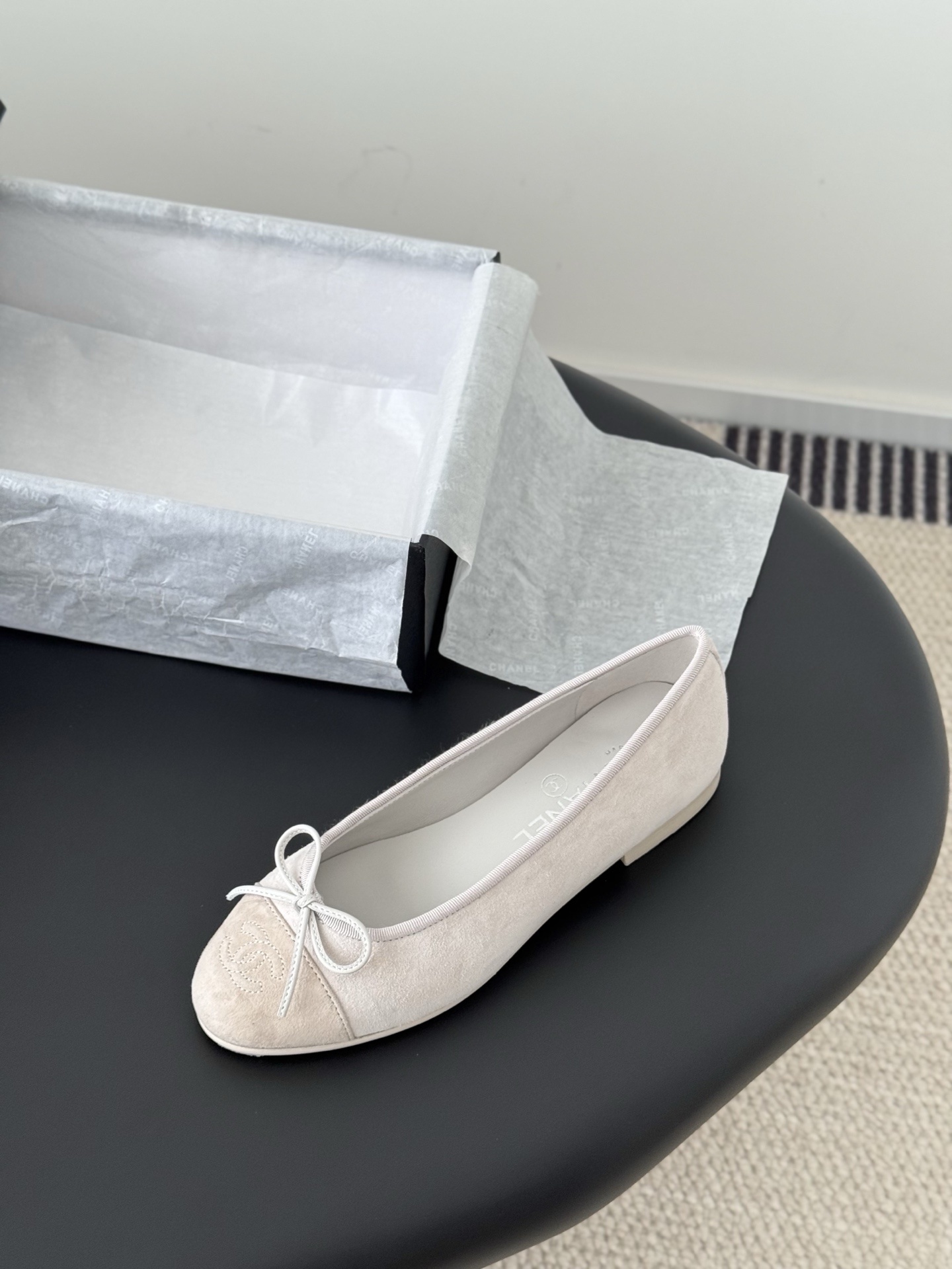 Pair of Chanel beige suede ballet flats with CC logo on the toe cap and white bow detail, placed next to a black Chanel box. - Hình 5