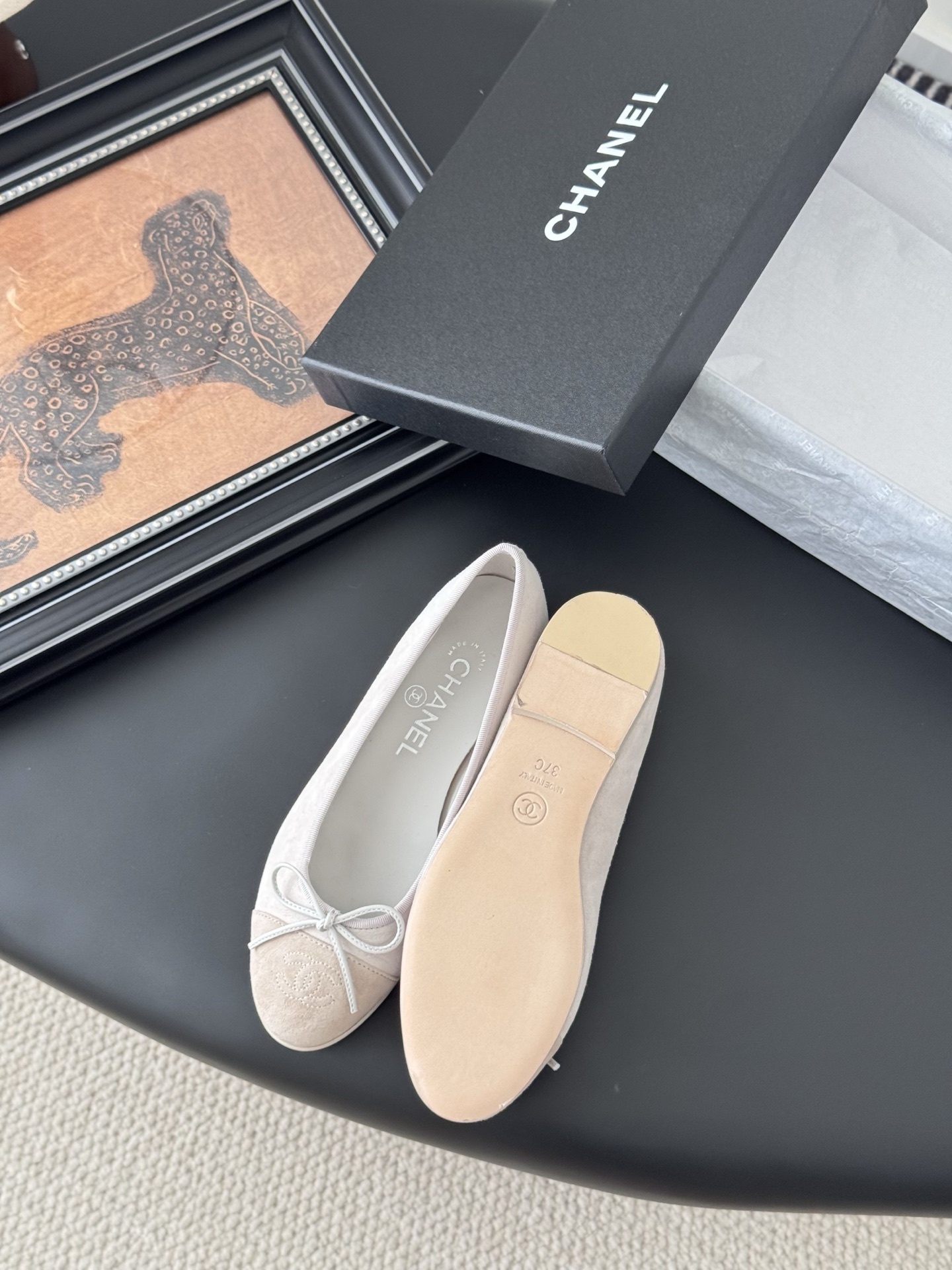 Pair of Chanel beige suede ballet flats with CC logo on the toe cap and white bow detail, placed next to a black Chanel box. - Hình 4