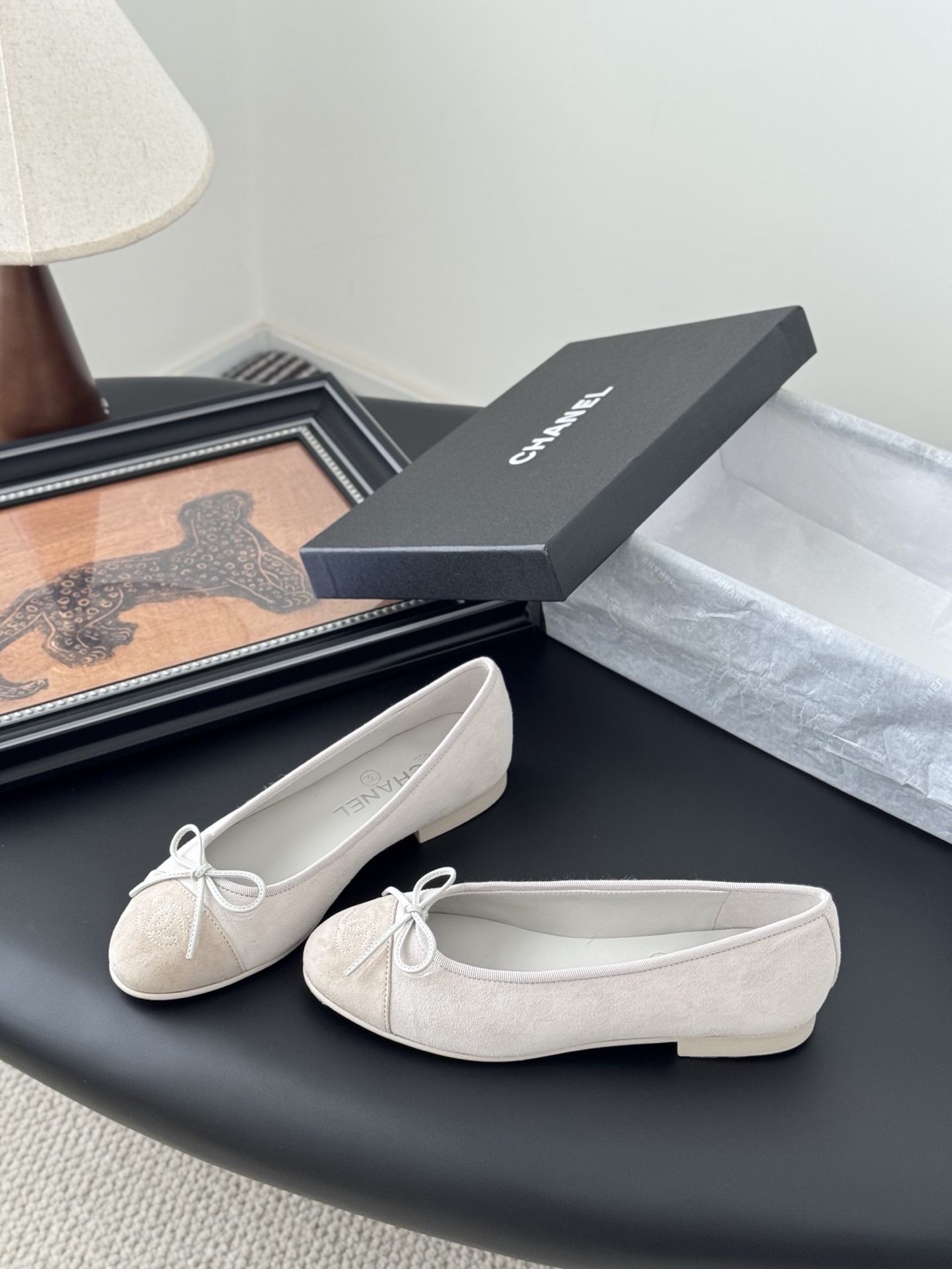 Pair of Chanel beige suede ballet flats with CC logo on the toe cap and white bow detail, placed next to a black Chanel box. - Hình 2