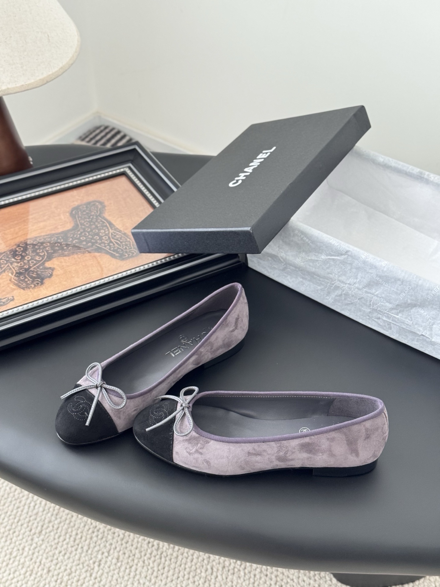 A pair of Chanel mauve suede ballet flats with black toe caps and bow detail next to an original Chanel box. - Hình 2