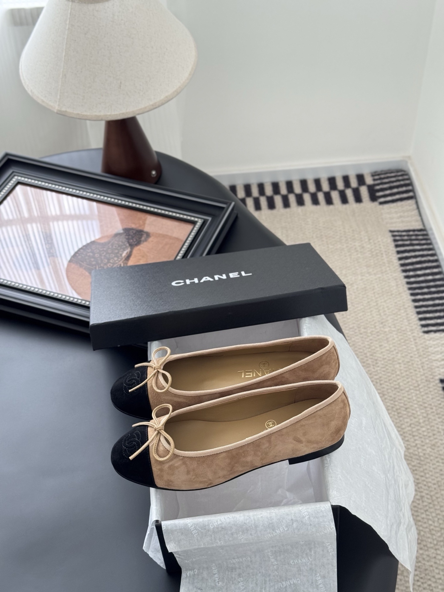 Pair of Chanel beige suede ballerina flats with black toe caps and bow detail next to a luxury black Chanel box. - Hình 9