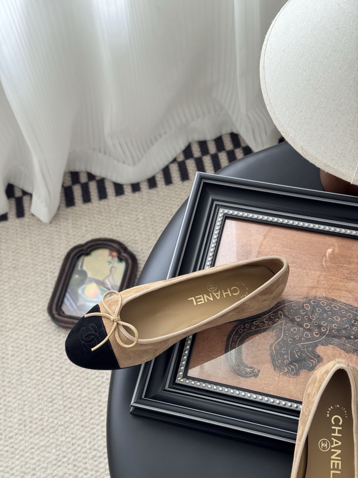 Pair of Chanel beige suede ballerina flats with black toe caps and bow detail next to a luxury black Chanel box. - Hình 8