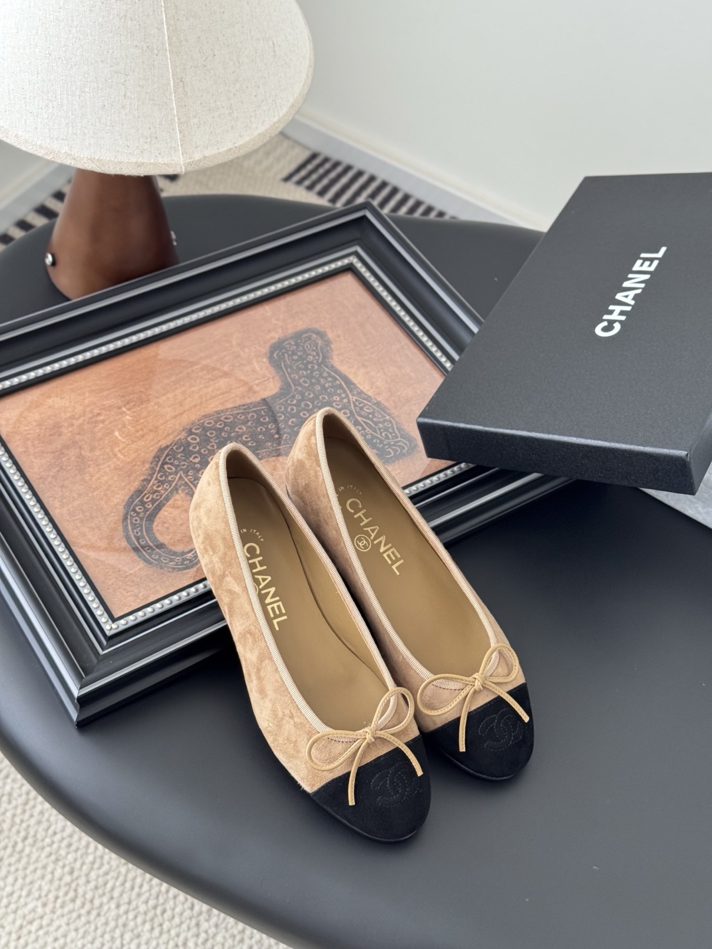 Pair of Chanel beige suede ballerina flats with black toe caps and bow detail next to a luxury black Chanel box. - Hình 7