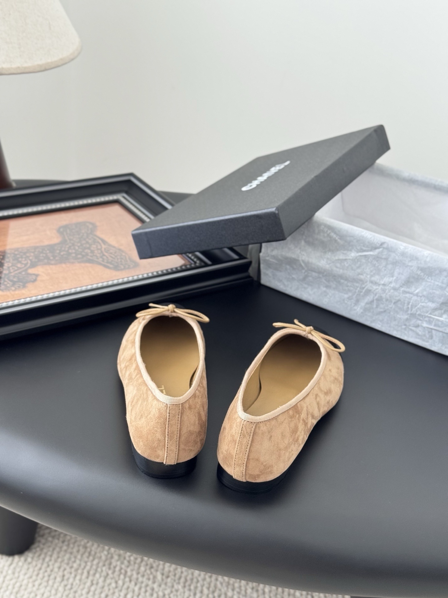 Pair of Chanel beige suede ballerina flats with black toe caps and bow detail next to a luxury black Chanel box. - Hình 6