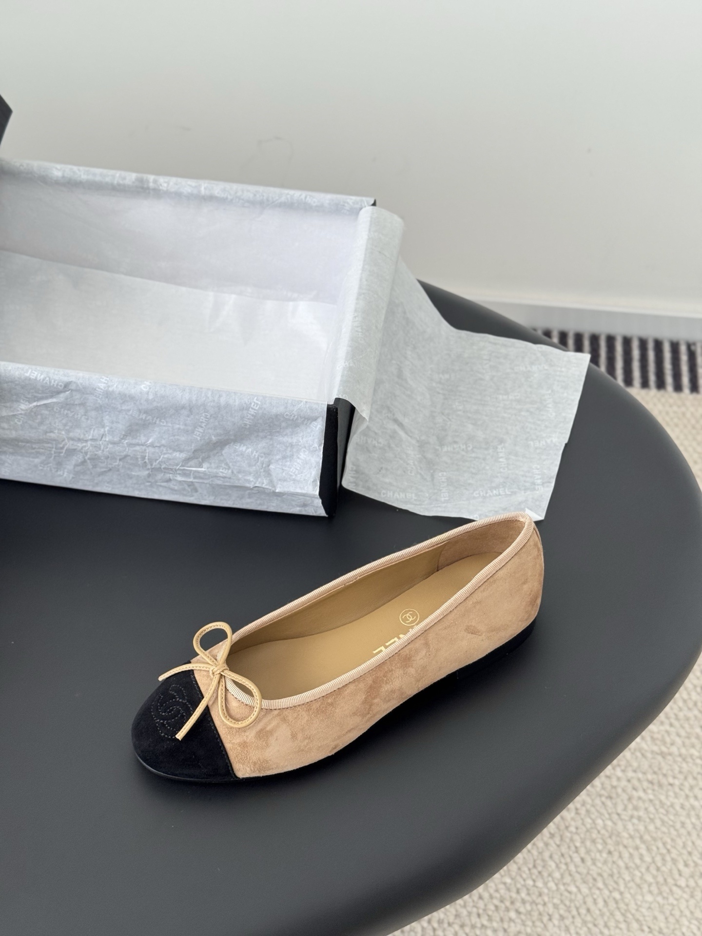 Pair of Chanel beige suede ballerina flats with black toe caps and bow detail next to a luxury black Chanel box. - Hình 5