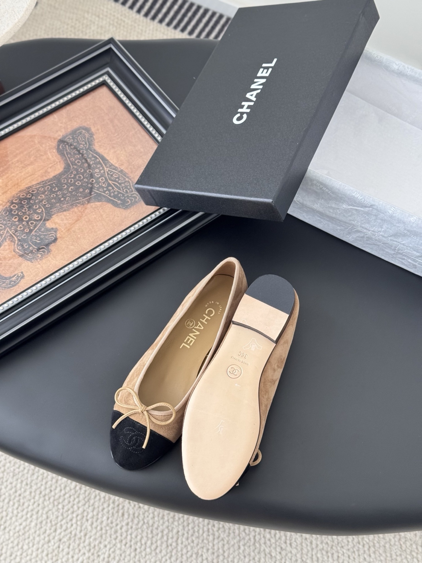 Pair of Chanel beige suede ballerina flats with black toe caps and bow detail next to a luxury black Chanel box. - Hình 4