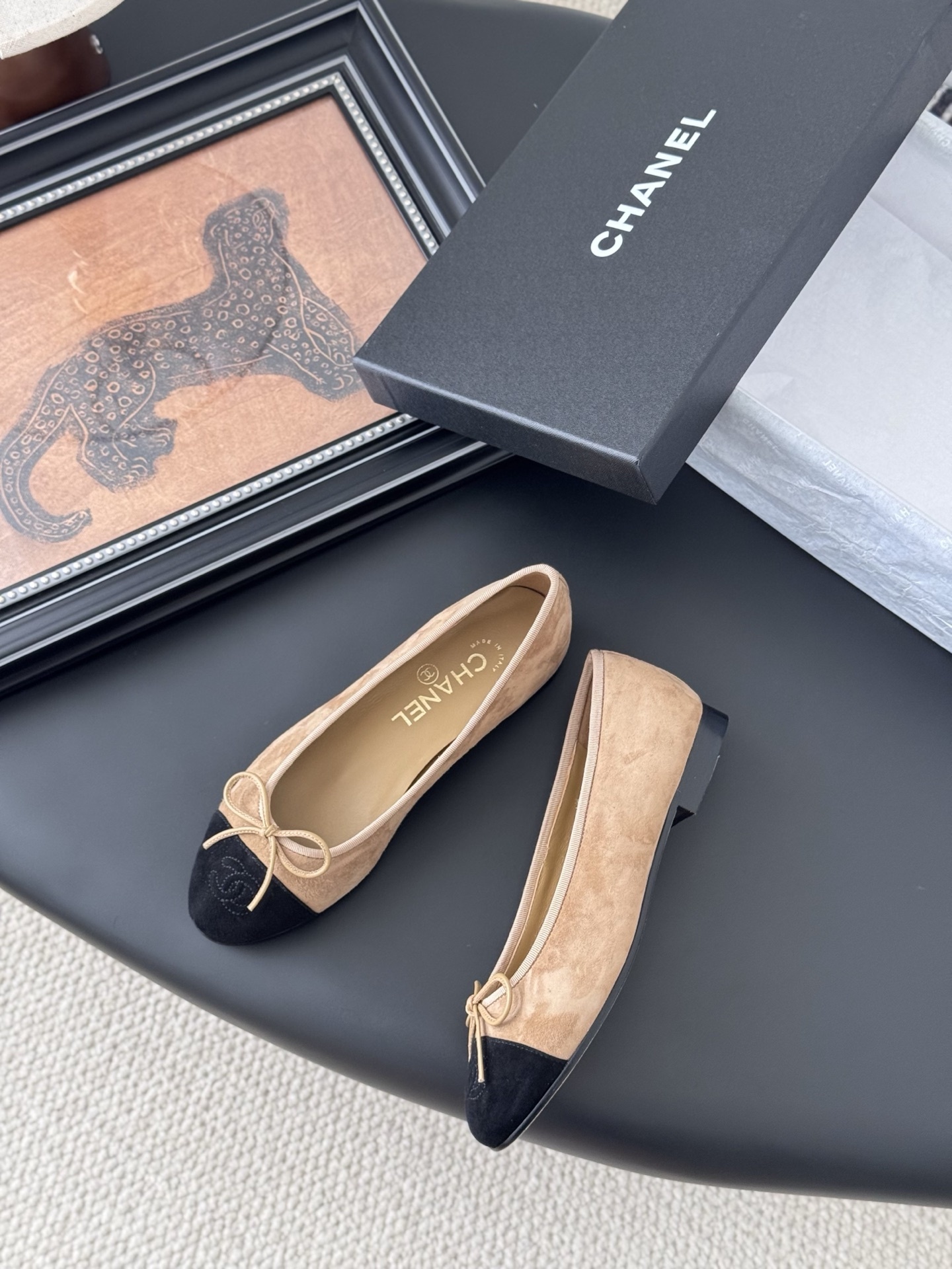 Pair of Chanel beige suede ballerina flats with black toe caps and bow detail next to a luxury black Chanel box. - Hình 3