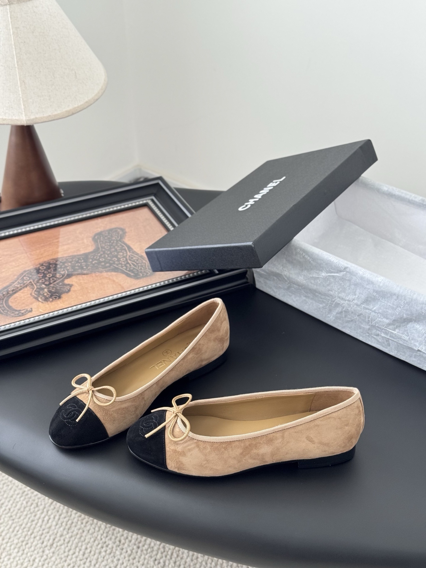 Pair of Chanel beige suede ballerina flats with black toe caps and bow detail next to a luxury black Chanel box. - Hình 2