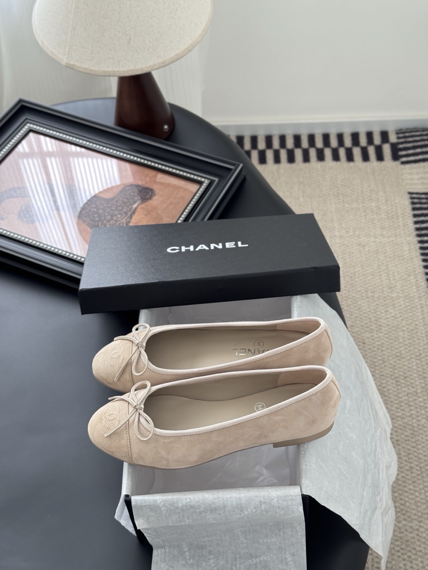 Pair of beige Chanel suede ballet flats with embossed CC logo and bow detail next to a black Chanel shoe box. - Hình 9