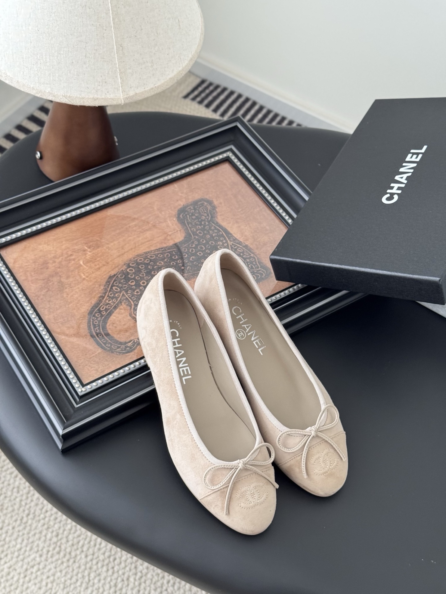 Pair of beige Chanel suede ballet flats with embossed CC logo and bow detail next to a black Chanel shoe box. - Hình 7