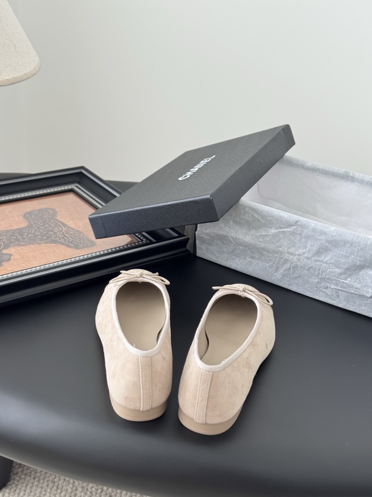 Pair of beige Chanel suede ballet flats with embossed CC logo and bow detail next to a black Chanel shoe box. - Hình 6