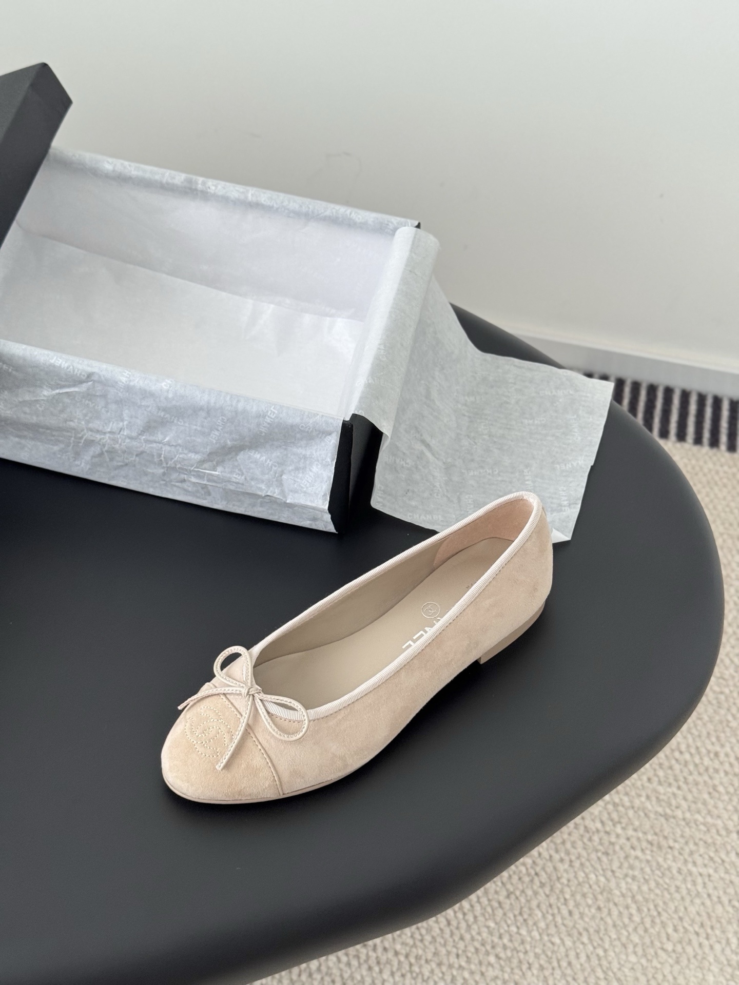 Pair of beige Chanel suede ballet flats with embossed CC logo and bow detail next to a black Chanel shoe box. - Hình 5